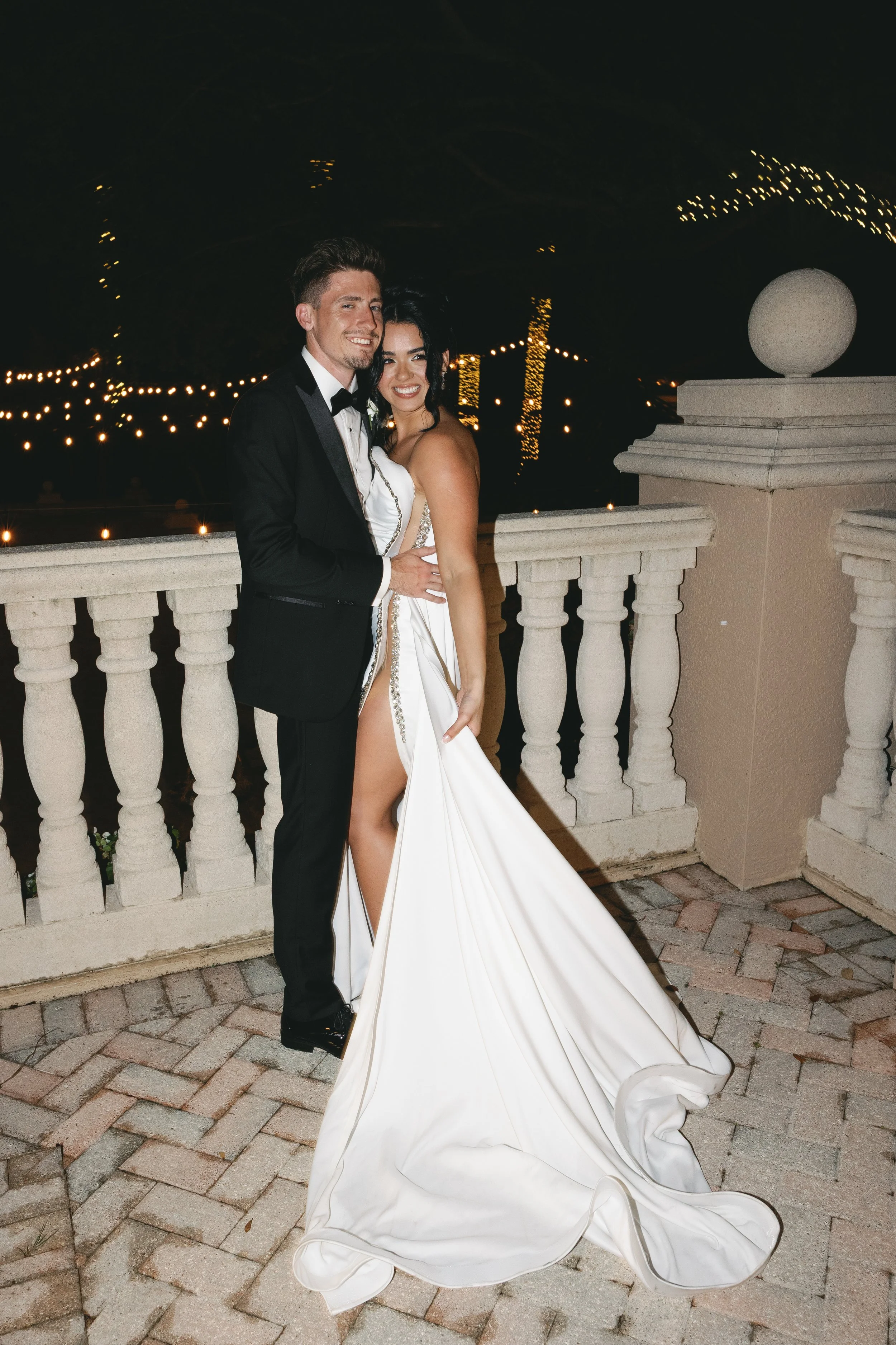 A couple dressed in wedding attire standing on a balcony at night, smiling at the camera. The man is wearing a black tuxedo and the woman is wearing a white wedding dress with a high slit, revealing her leg. The background features string lights and 