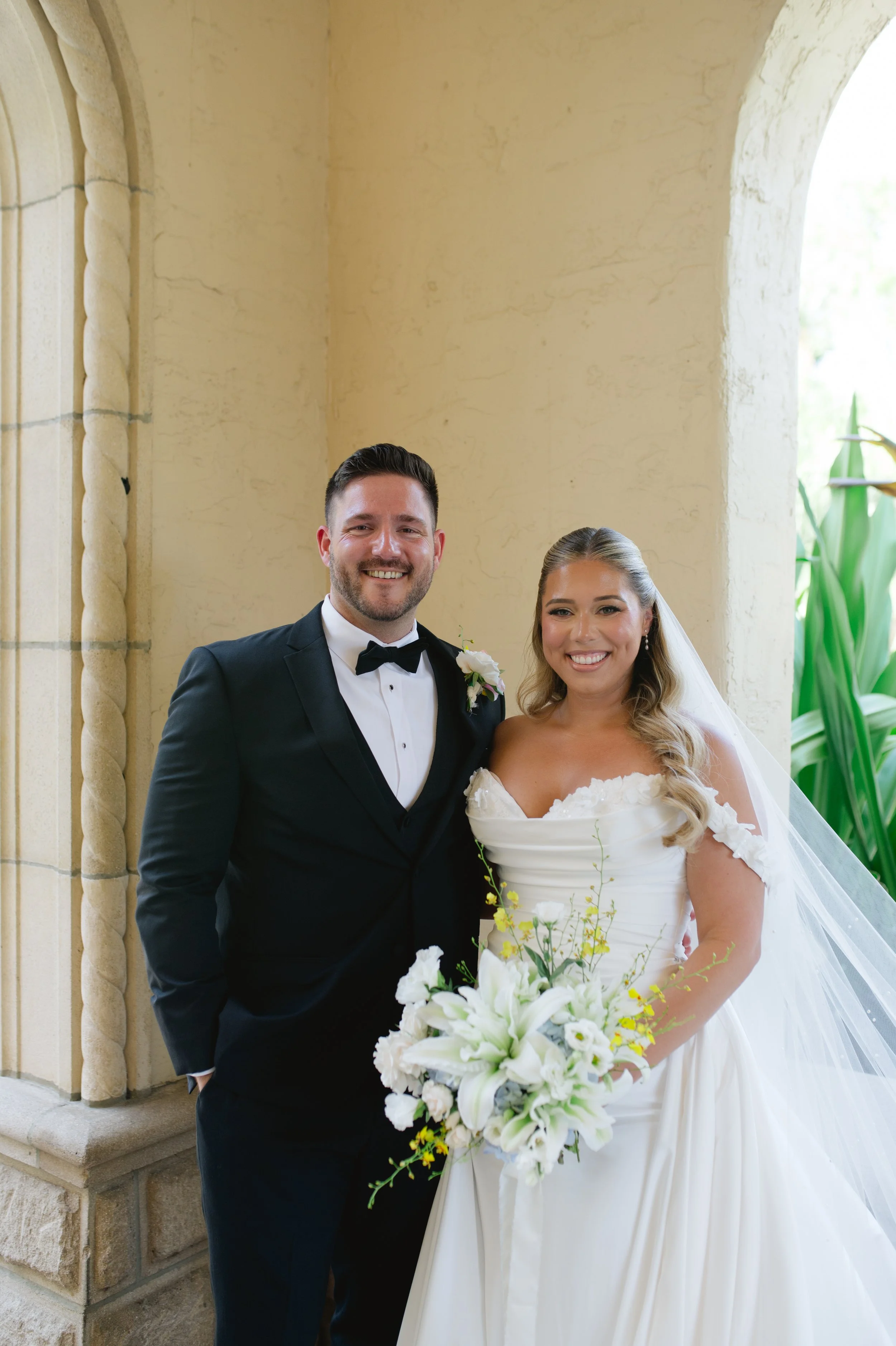 A bride and groom posing together at their wedding, standing under an archway with stone columns. The groom is in a tuxedo with a bowtie, and the bride is in a white wedding dress holding a bouquet of white flowers with greenery. Both are smiling.