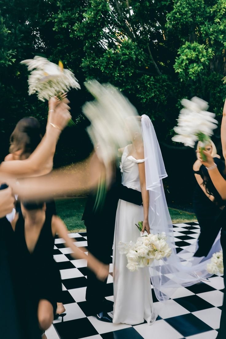 A bride holding a bouquet of white flowers standing on a checkered black and white floor, surrounded by people throwing white flower petals in the air, with greenery in the background.