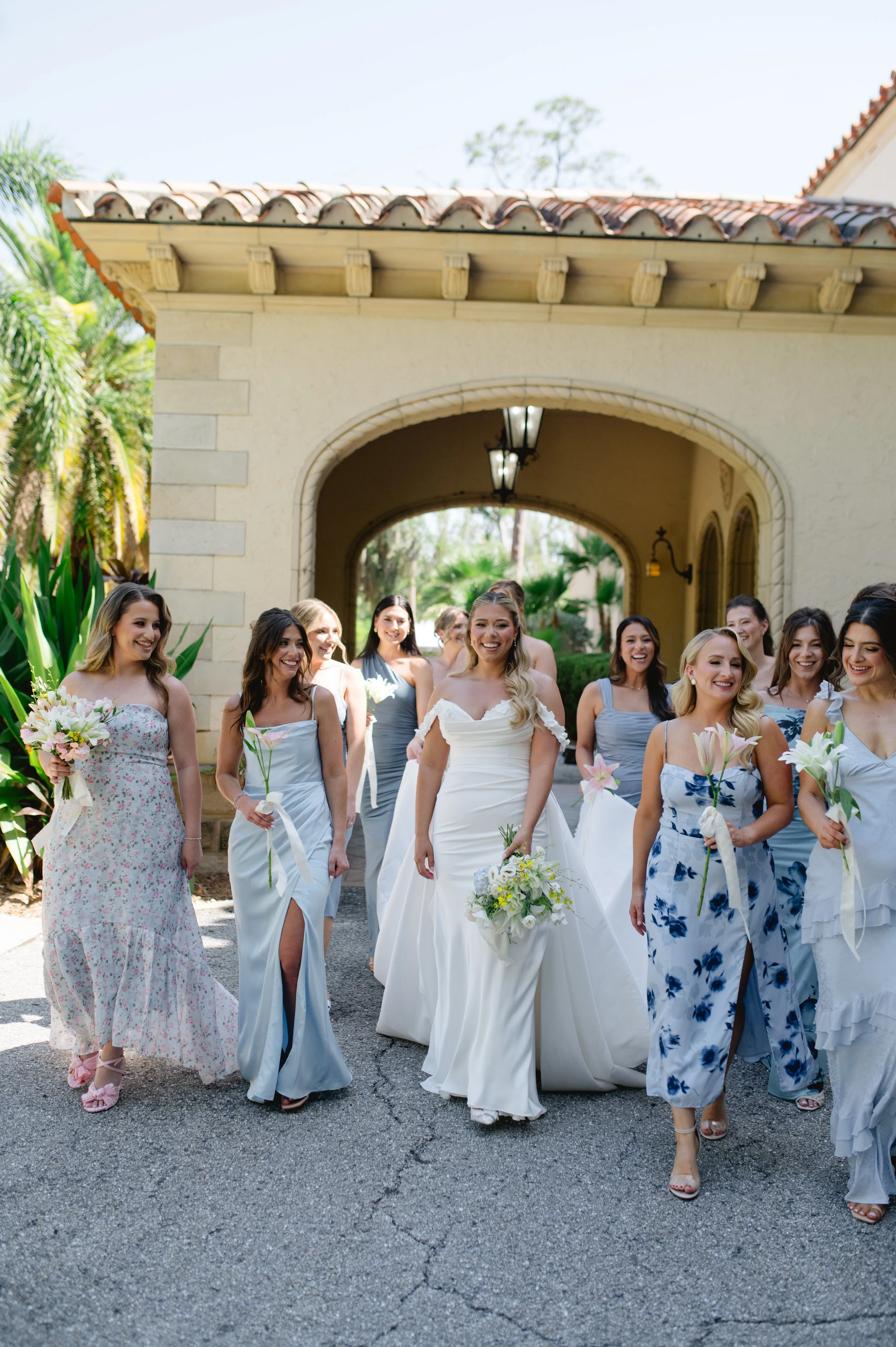 A group of women in elegant dresses, including a bride in a white wedding gown, walking outside a building with a stone archway, surrounded by lush greenery and palm trees, smiling and holding flowers.