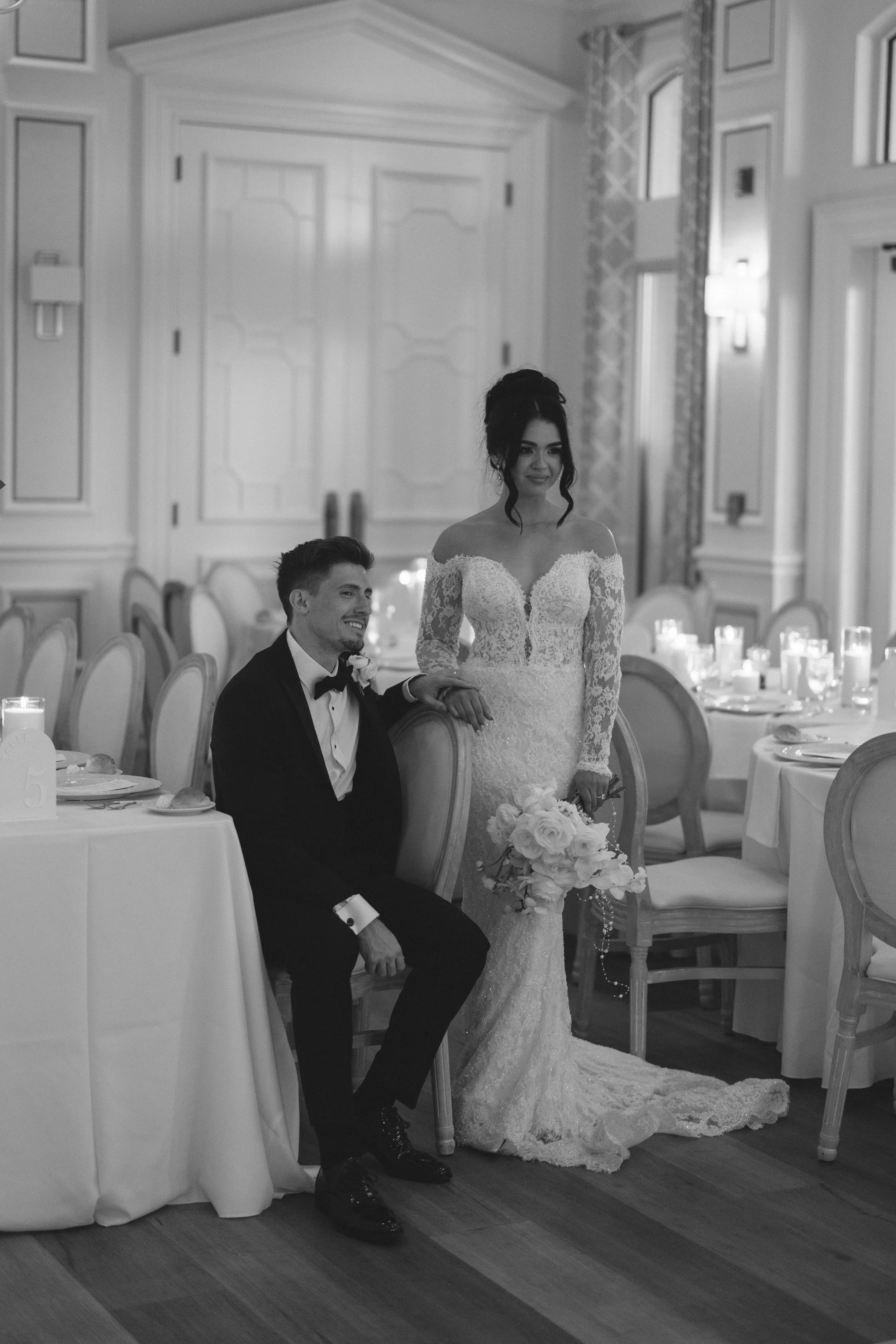 Black and white photo of a bride and groom at a wedding reception. The groom is sitting in a chair smiling, wearing a tuxedo with a bowtie. The bride is standing beside him, holding his hand, wearing an off-the-shoulder lace wedding gown and holding 