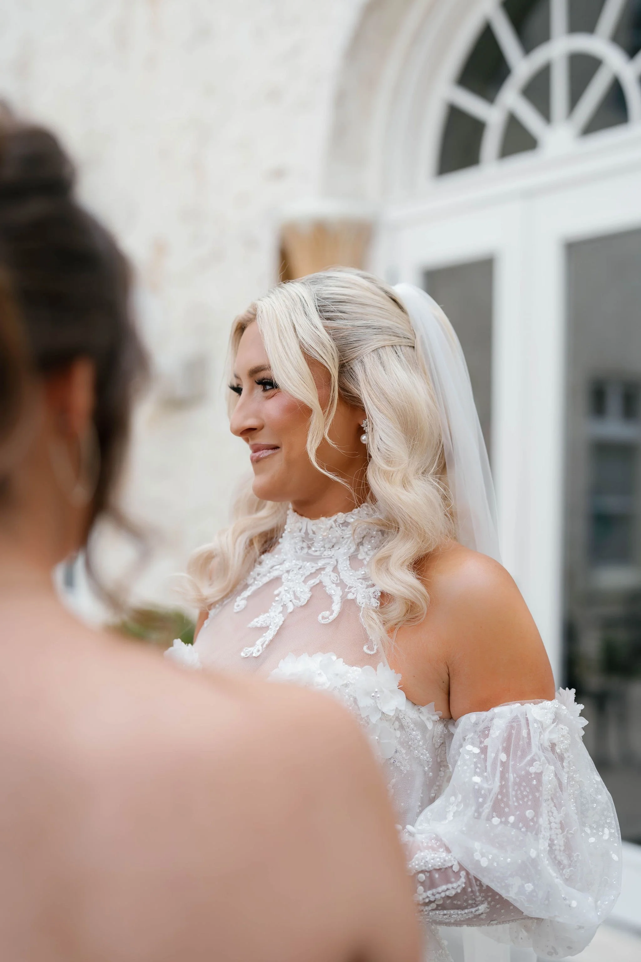 A bride with long, blonde, curly hair wearing a white lace wedding gown and veil, smiling as she looks at someone off-camera in an indoor setting with arched windows.