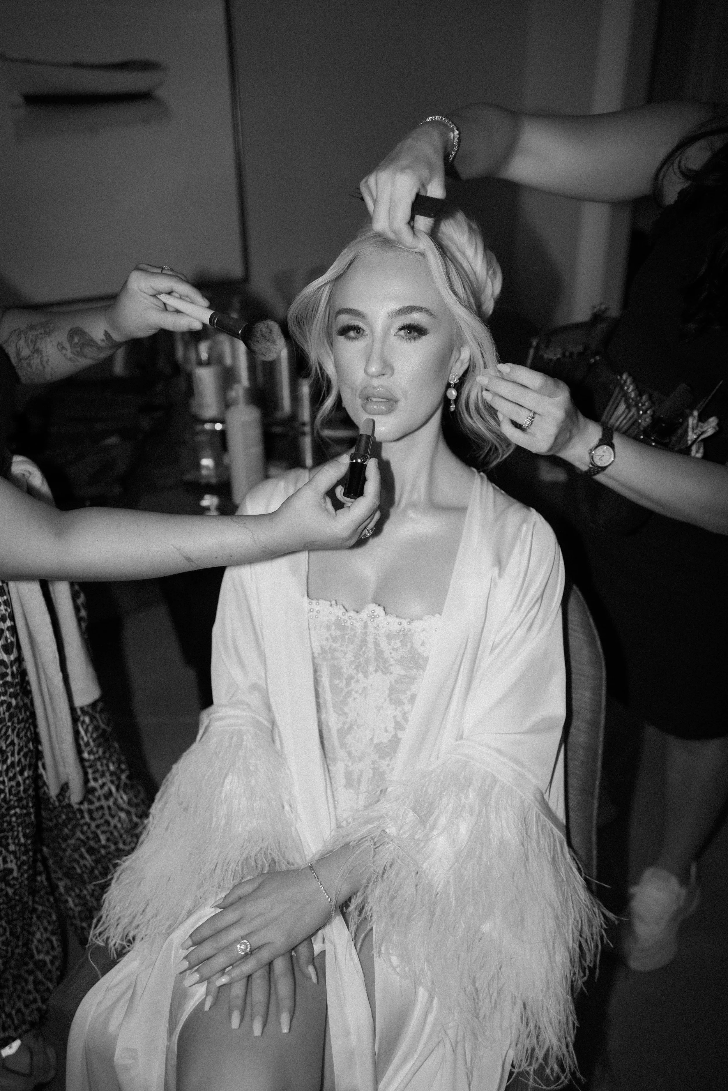 A woman with blonde hair and pearl earrings getting makeup done by several artists; one is applying lipstick while another applies eye makeup; the woman wears a light-colored dress with lace and feather details, and her hands are resting in her lap.