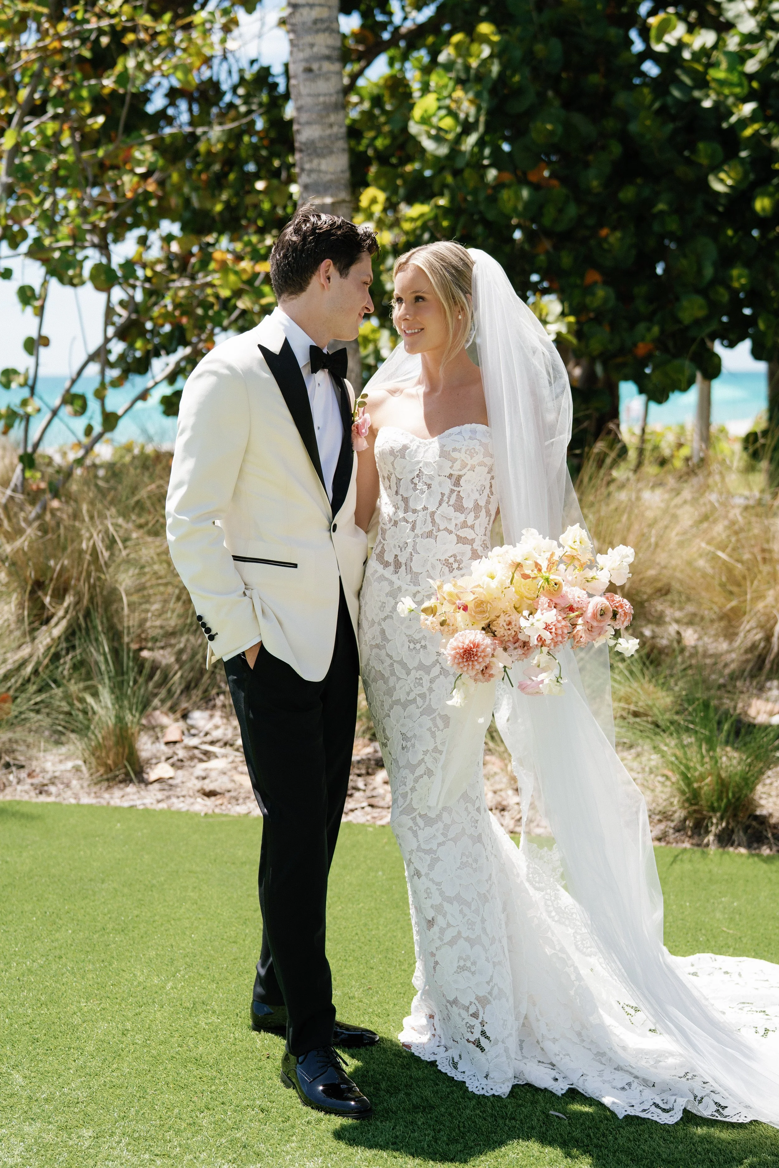 A bride and groom standing outdoors on a lush green lawn, facing each other, with trees and a clear sky in the background. The bride is wearing a strapless lace wedding gown with a long veil and holding a bouquet of pink and white flowers. The groom 
