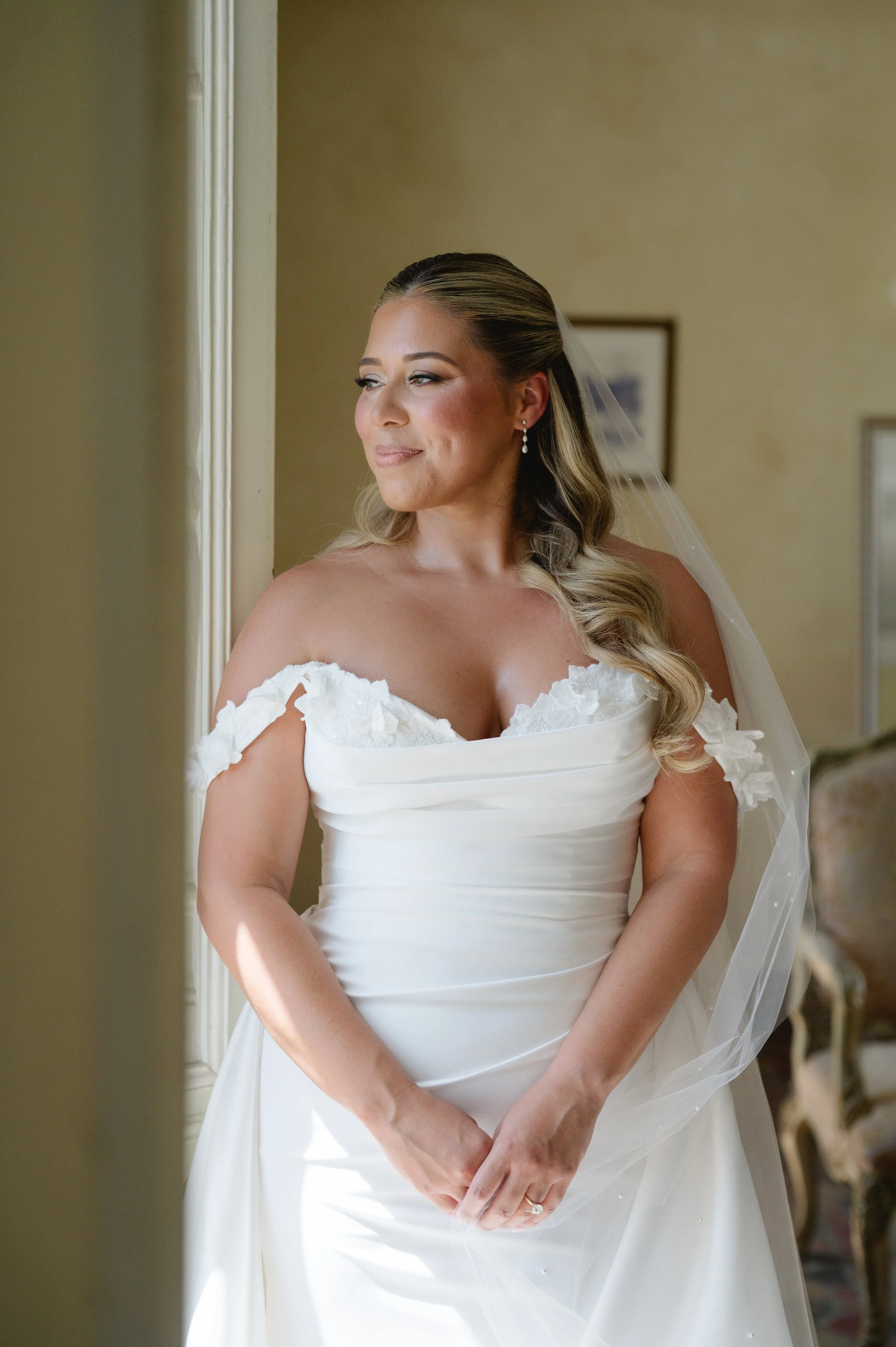 A bride with light skin, styled blonde hair, wearing pearl earrings, in a white wedding dress with off-the-shoulder lace details, standing indoors, gazing softly to her left, with a veil and a delicate ring on her finger.