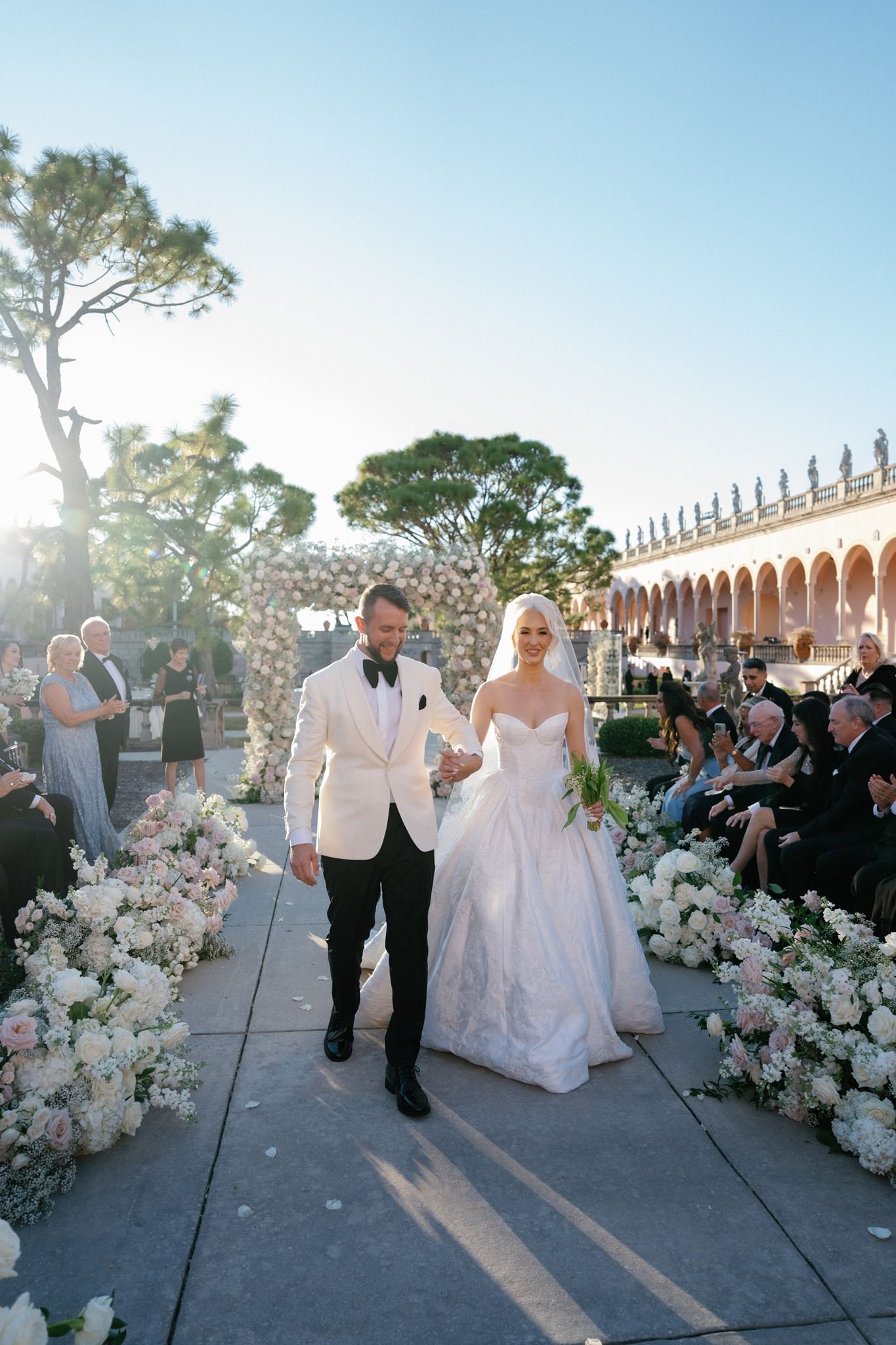 A newly married couple smiling as they walk down an aisle decorated with white and pale pink flowers at an outdoor wedding ceremony during sunset. The groom is wearing a white tuxedo with a black bow tie, and the bride is in a white wedding gown hold