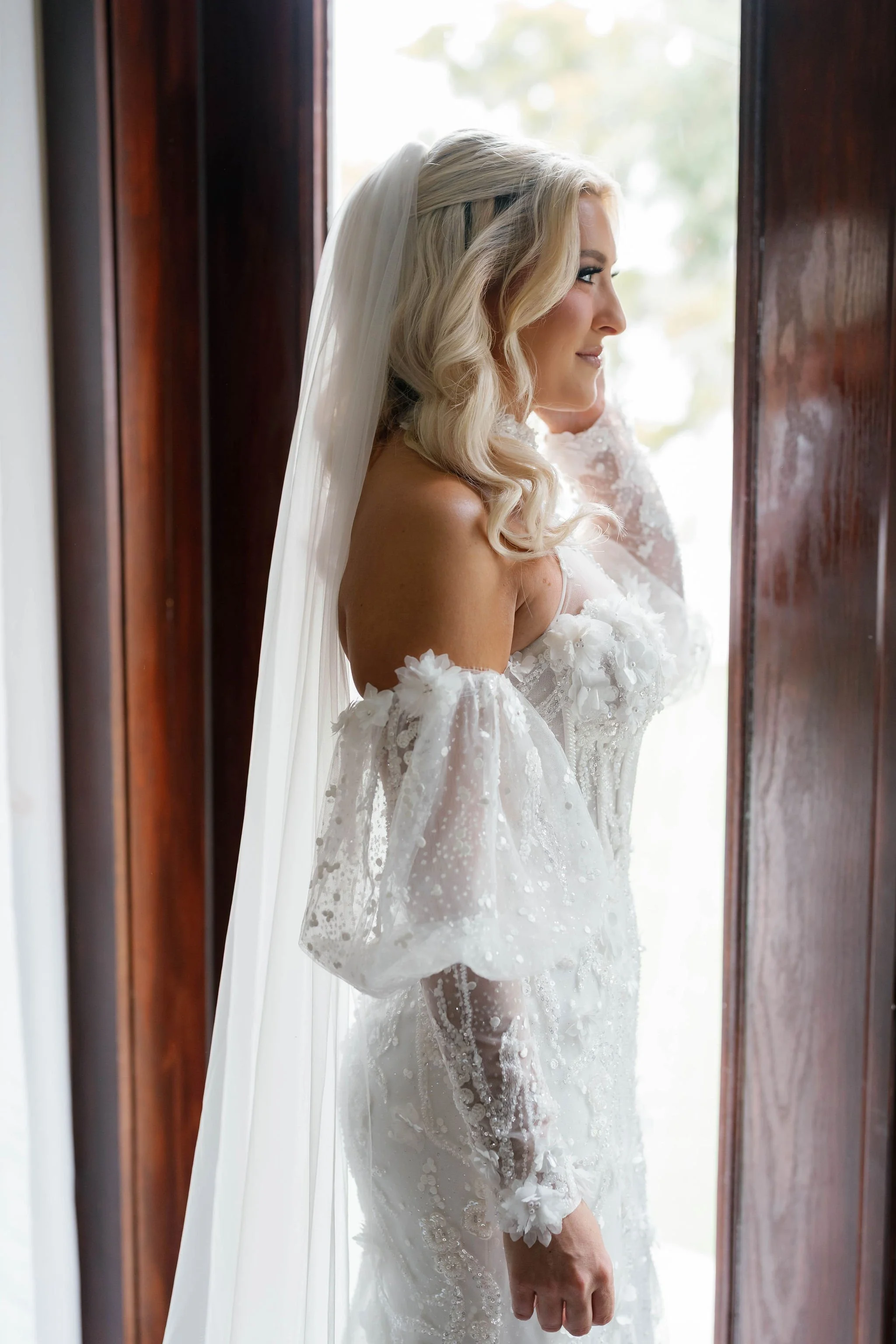 A bride standing by a window, smiling, in a wedding dress with lace detail and a veil.