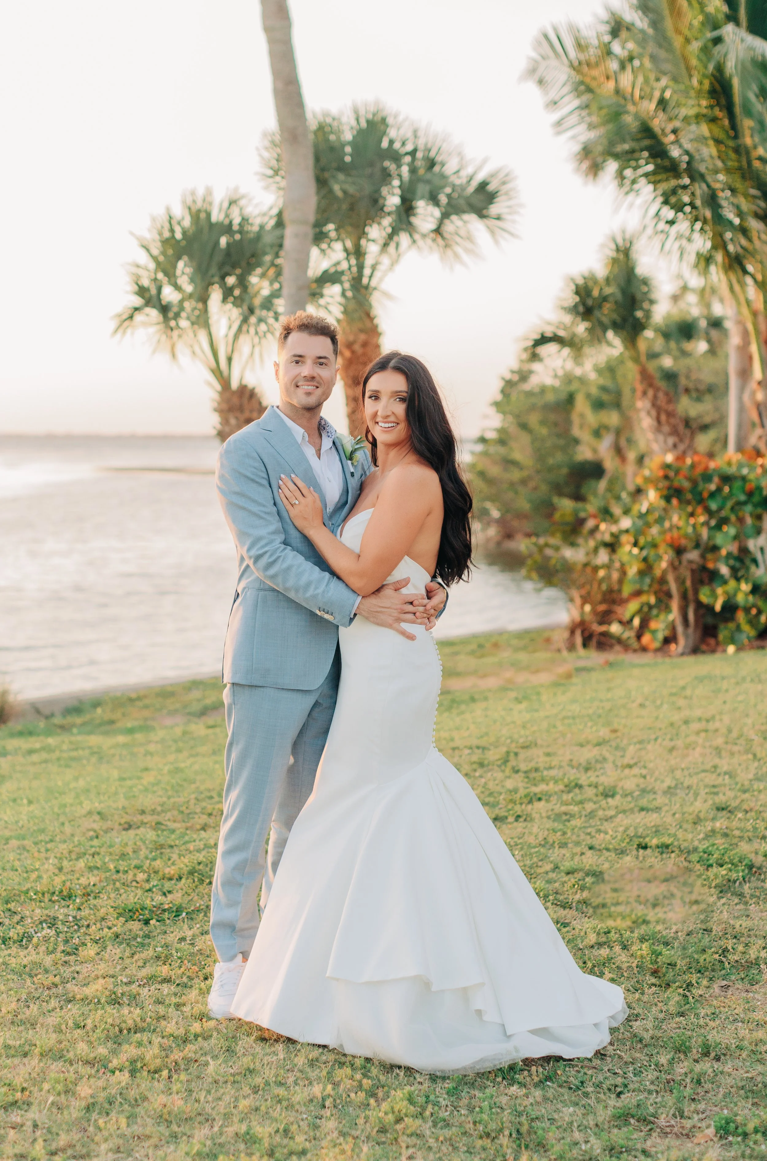 A newlywed couple standing outdoors on a grassy area near water, with palm trees in the background during sunset. The groom wears a light gray suit and the bride wears a strapless white wedding gown. They are embracing and smiling at the camera.