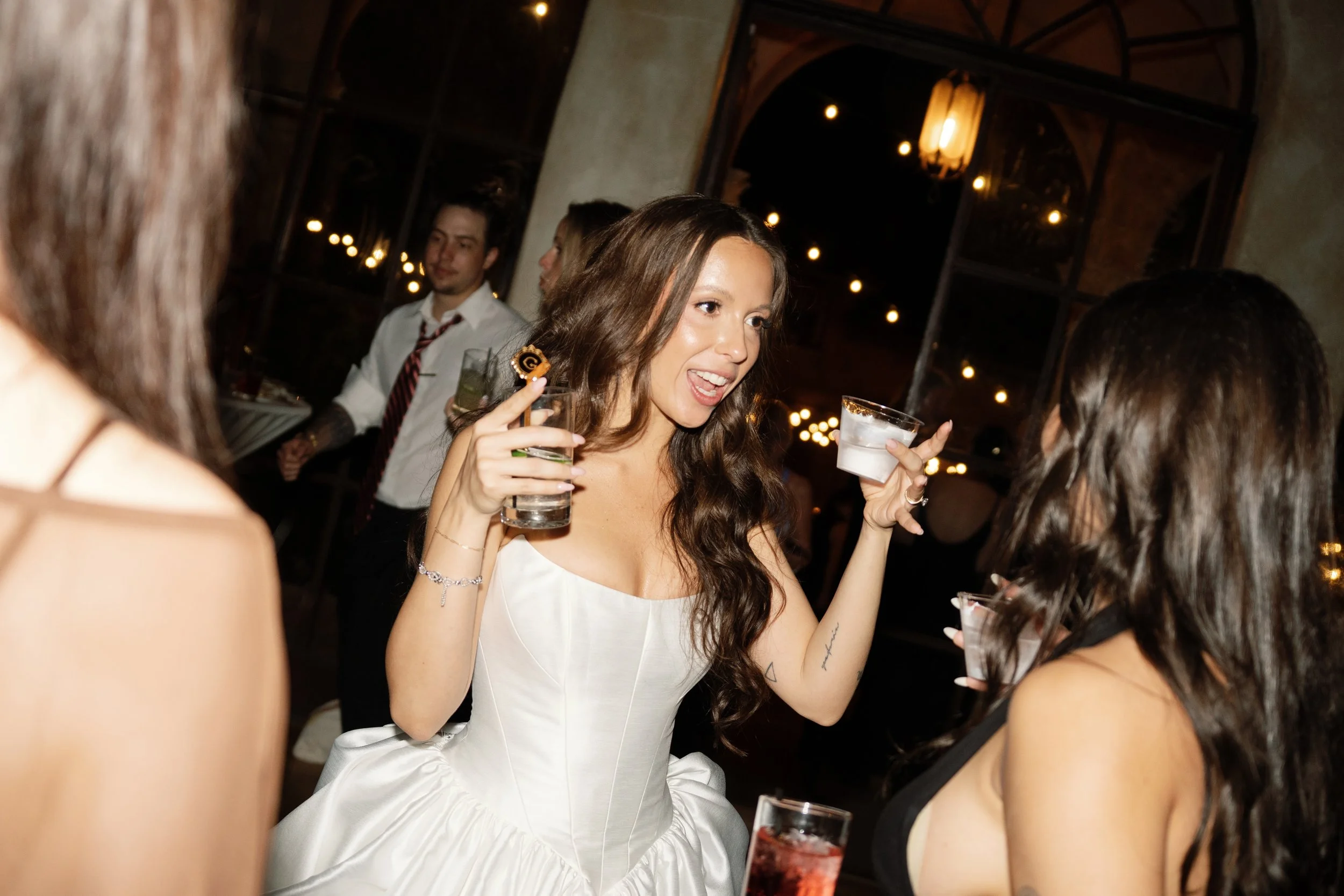 A woman in a white dress holding a clear drink, smiling and talking to another woman at a party or celebration in a dimly lit venue with string lights in the background.