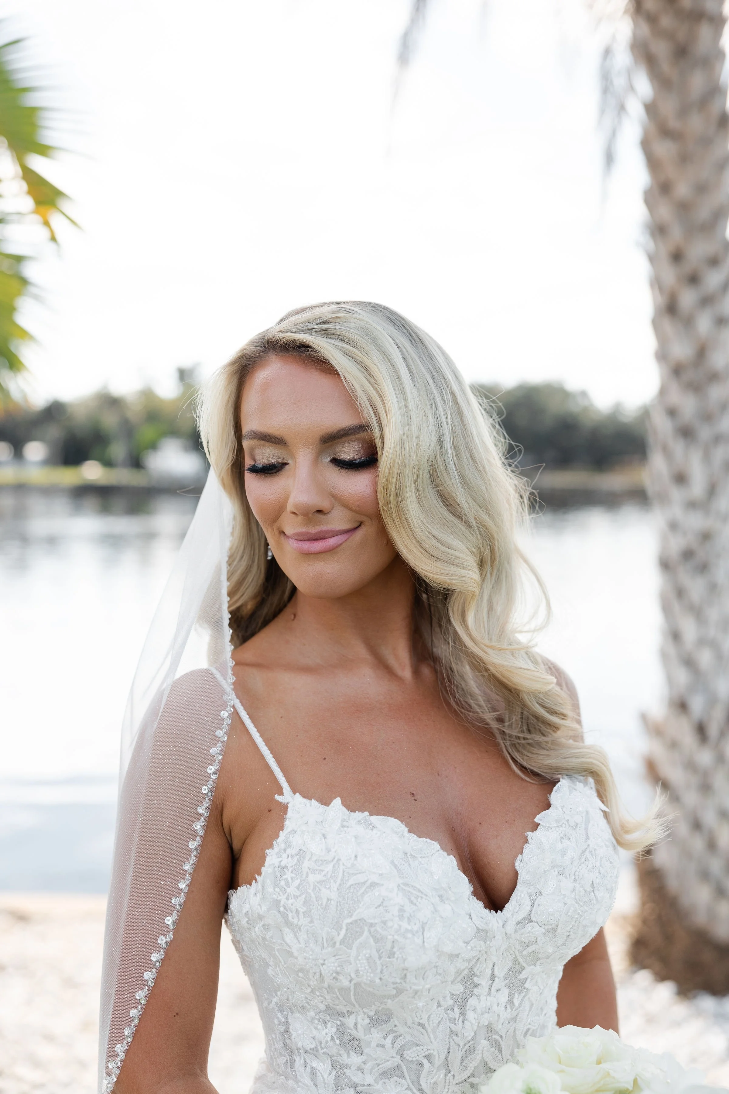 A bride with blonde hair and light skin, wearing a white lace wedding dress with a sheer sleeve, stands outdoors near a lake with palm trees in the background, holding a bouquet of white flowers.