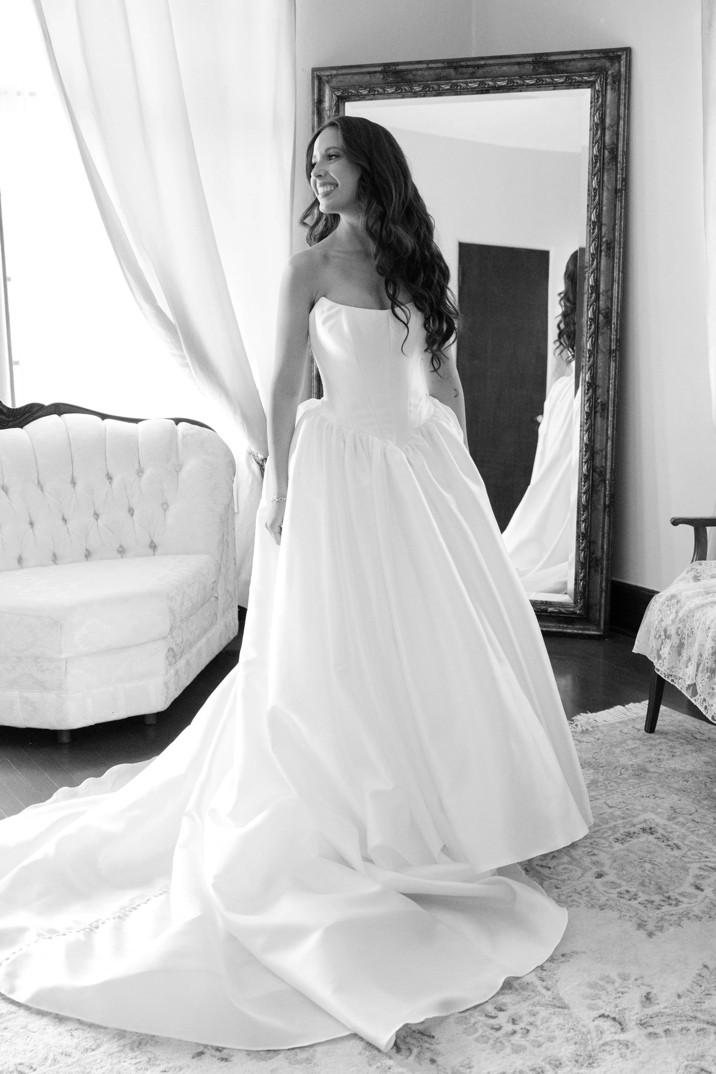 A woman in a wedding gown smiling while looking in a mirror in a room with vintage furniture and a curtained window.