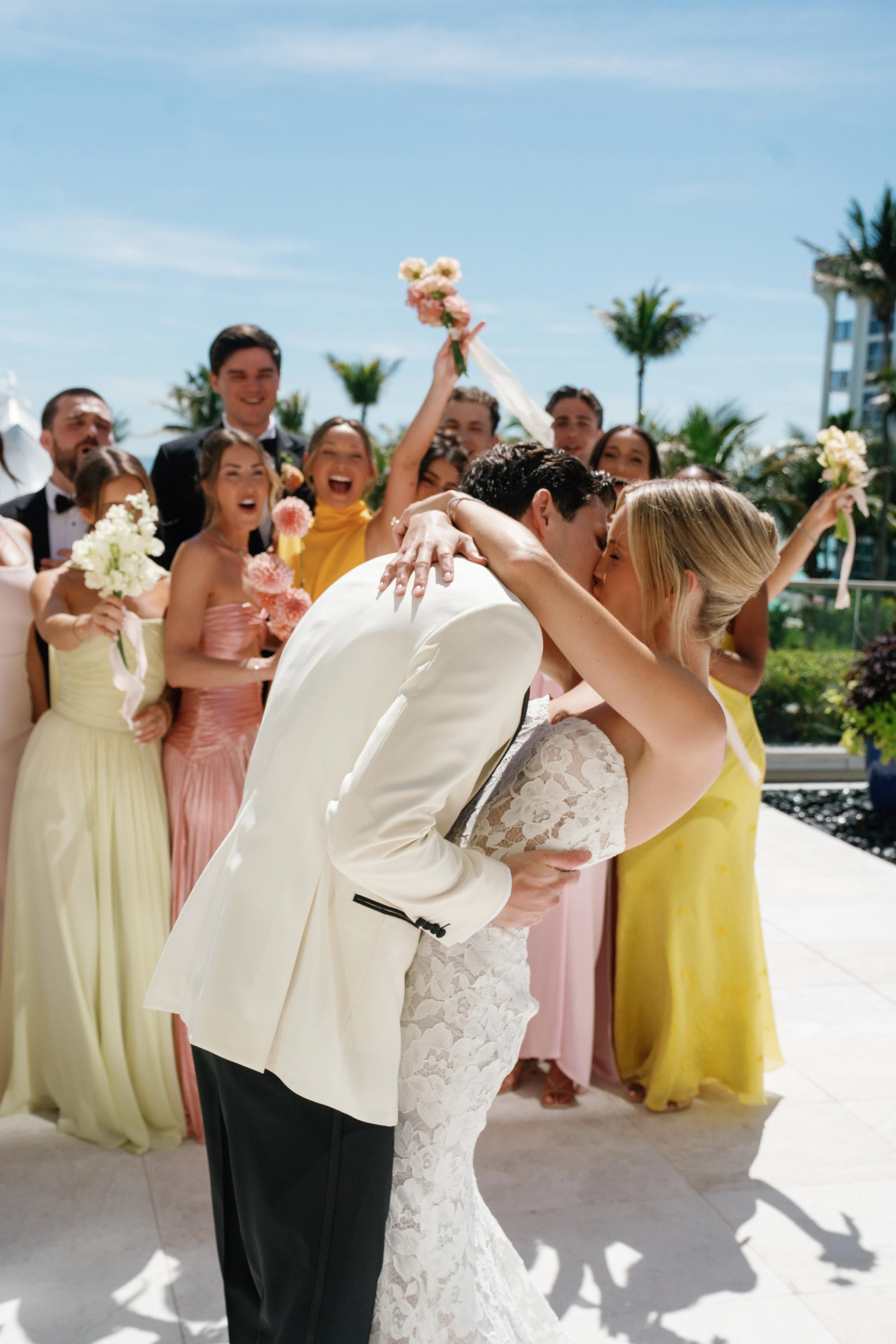 A bride and groom kiss while a group of wedding guests celebrate behind them, outdoors on a sunny day with sky and palm trees in the background.