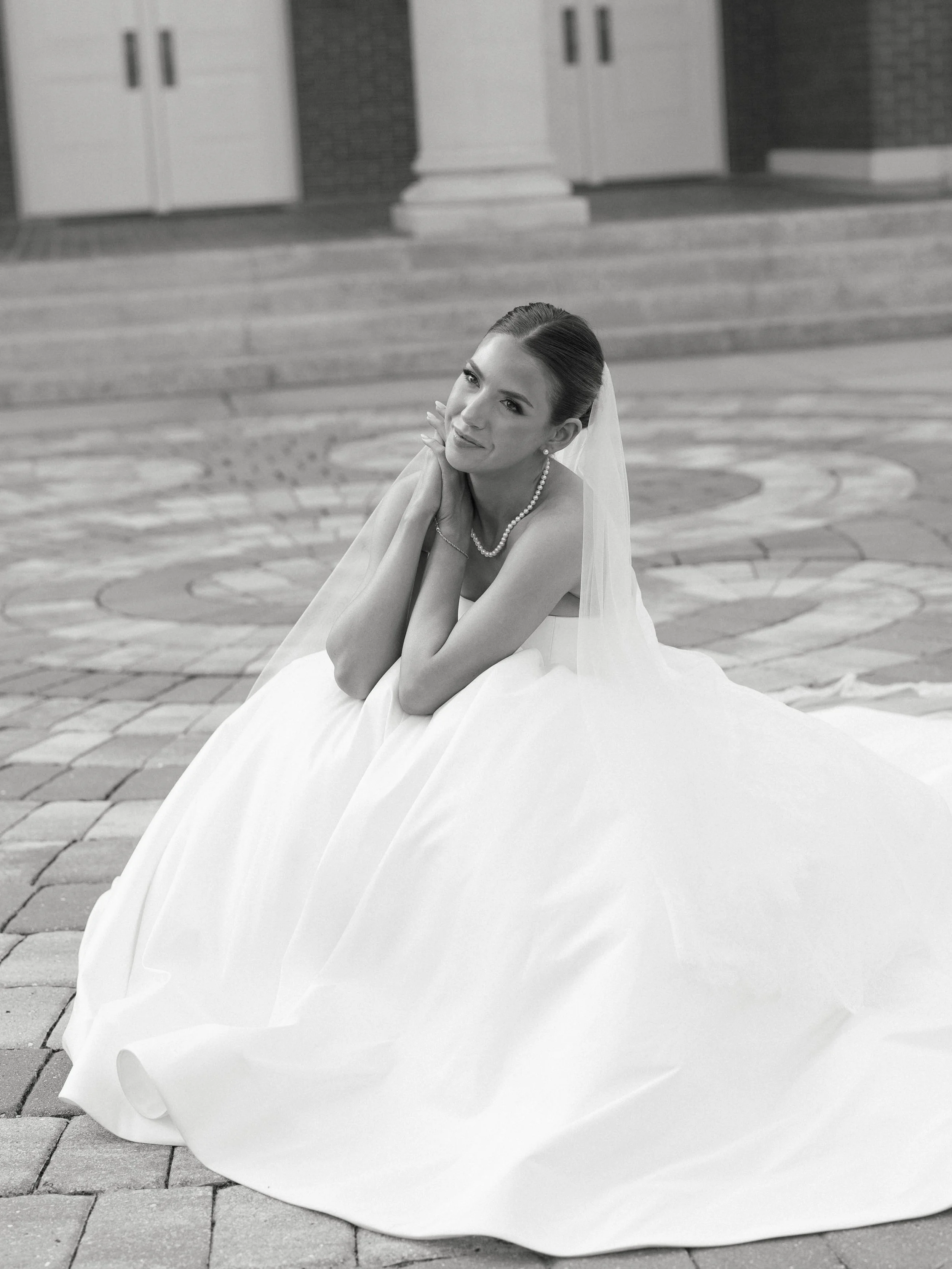 Black and white photo of a bride sitting on the steps of a building with large pavers, wearing a wedding dress, pearls, and a veil, with a calm expression.