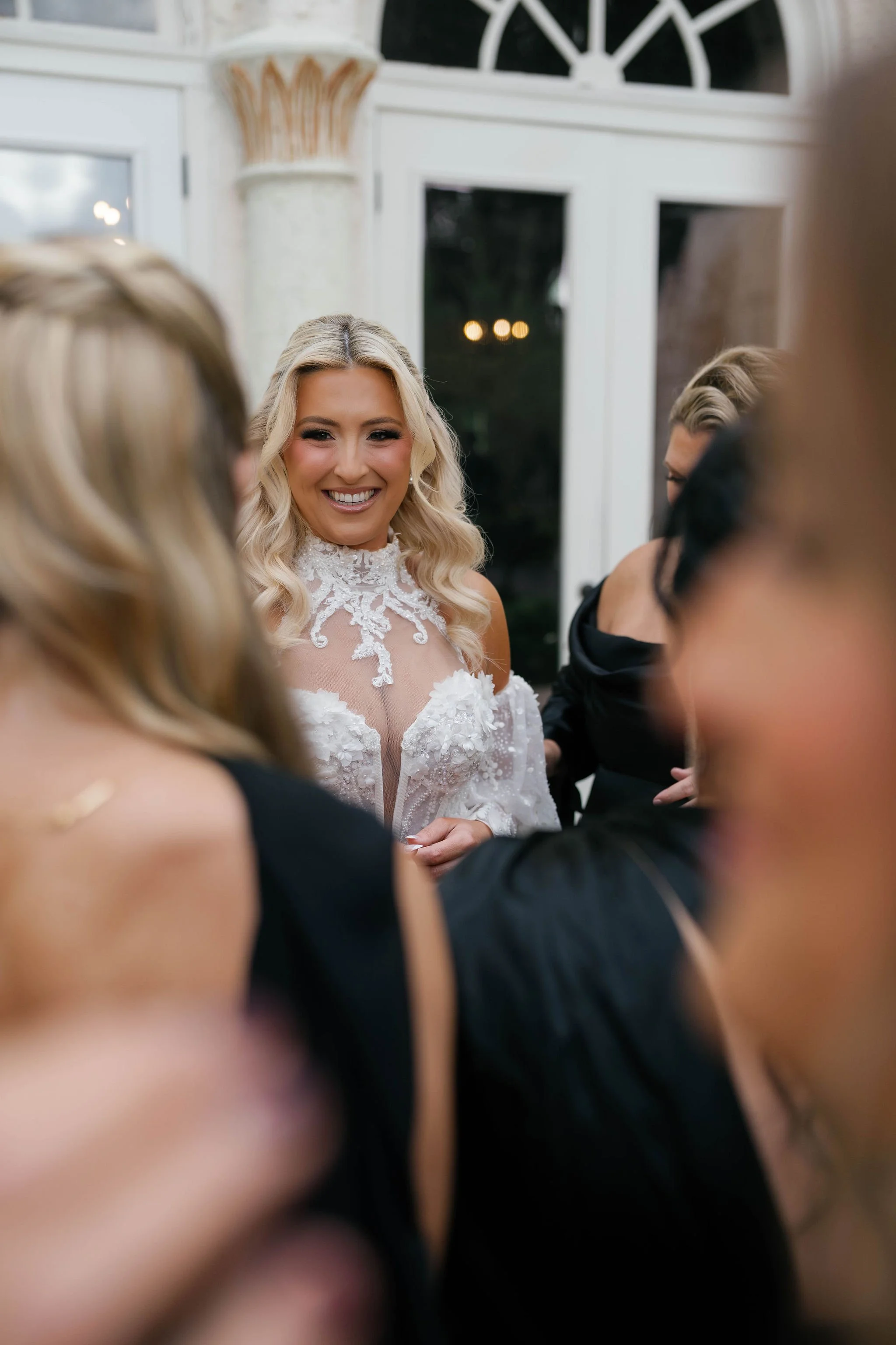 A group of women in elegant black dresses gathered around a smiling blonde woman in a wedding dress with lace details at a formal event or celebration indoors near a window.