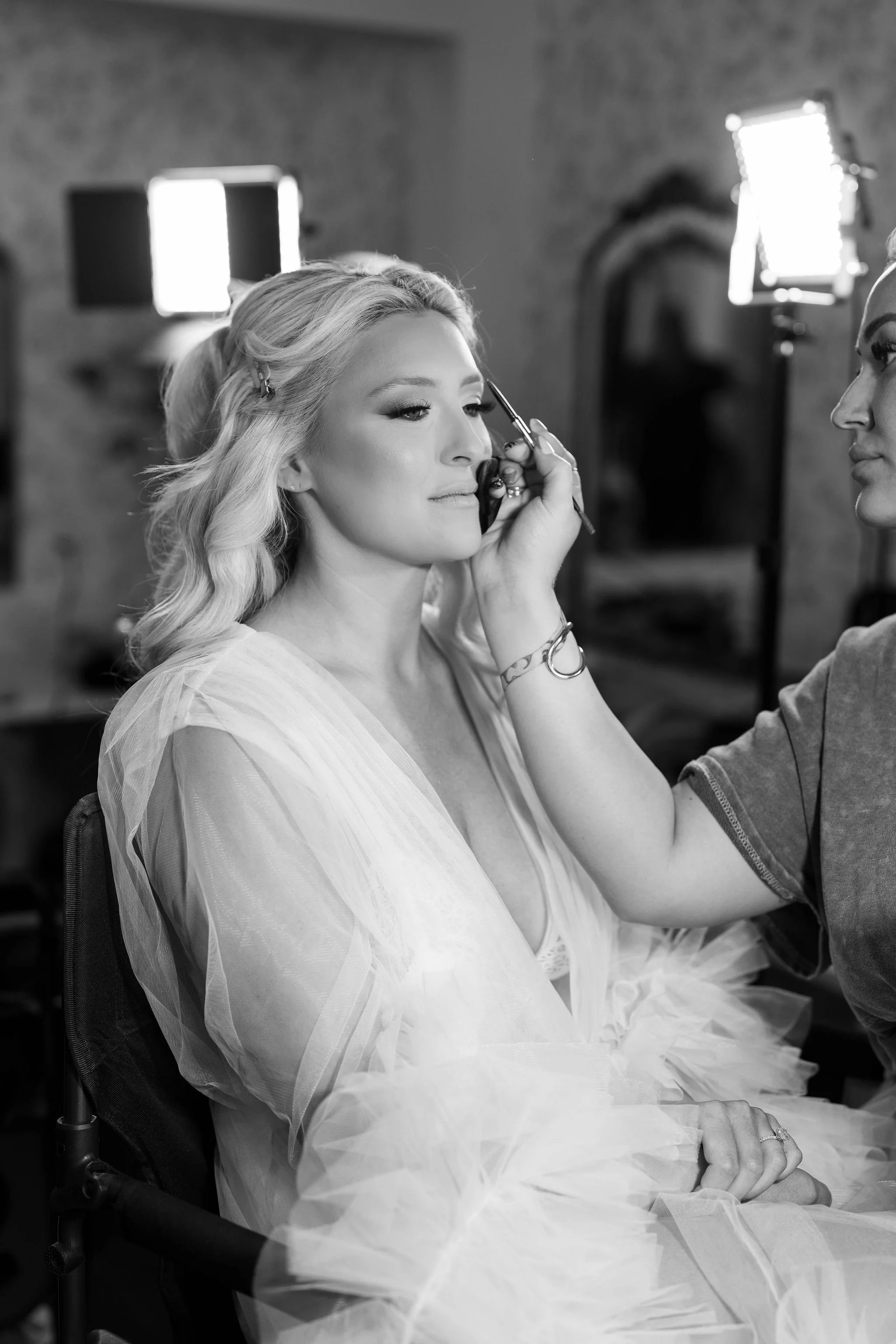 A woman with blonde hair and light makeup getting her eyebrows done by a stylist who is holding an eyebrow pencil. She is seated and wearing a sheer, flowing dress with tulle fabric on her lap. The background shows studio lighting equipment and a bri