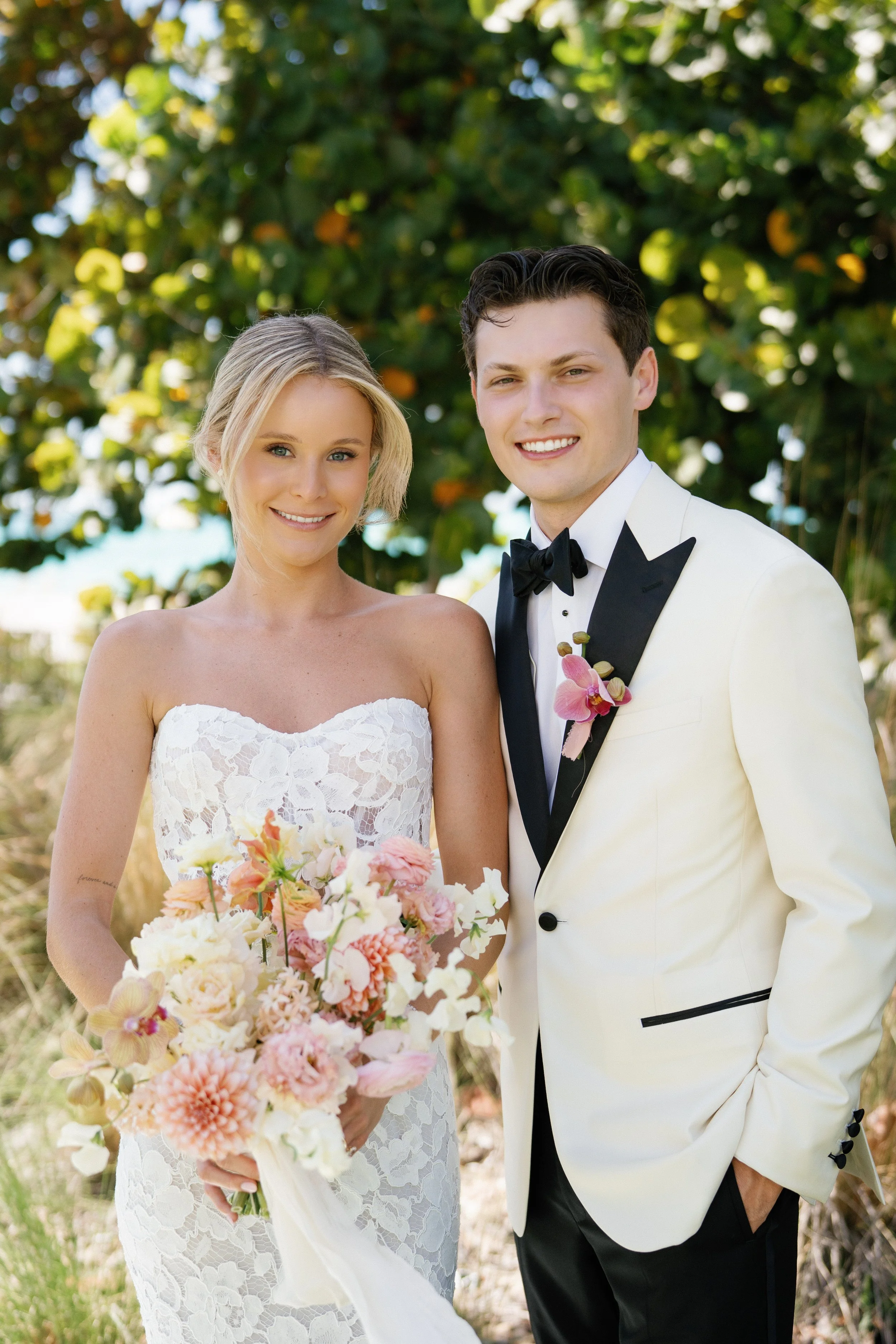 A smiling couple on their wedding day outdoors, the bride holding a bouquet of pink, peach, and white flowers, standing next to the groom in a cream tuxedo with a black bow tie.