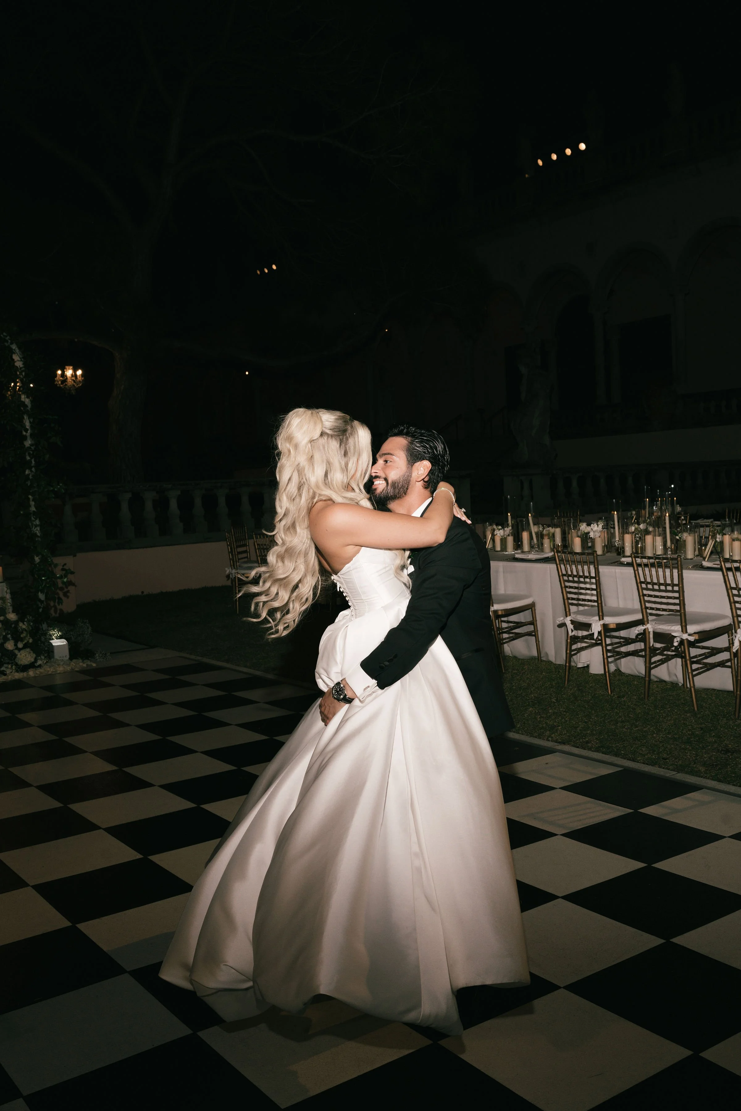 A newlywed couple sharing a dance at their wedding reception. The bride is in a white wedding gown and the groom is in a black suit, embracing each other with joyful expressions. In the background, tables are set with candles and floral centerpieces,
