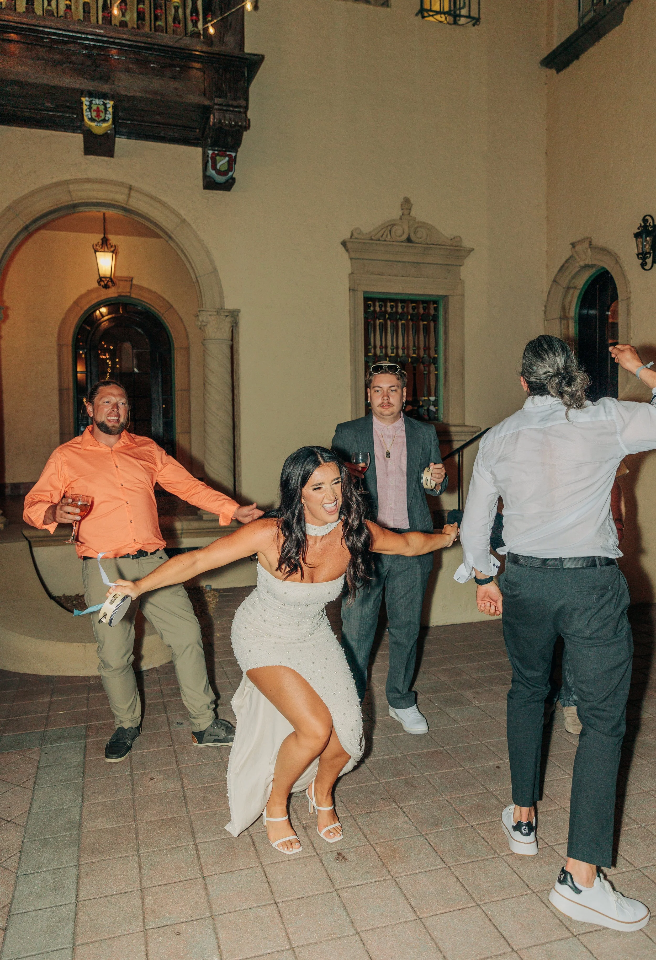 A group of people dancing and celebrating outdoors at night. The woman in the white dress is the focal point, smiling and dancing, with men around her also enjoying the moment.