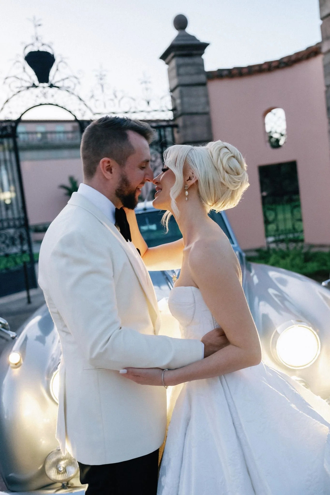 A bride and groom embracing outdoors near a vintage car decorated with lights, with a gate and wall in the background, during sunset or evening.