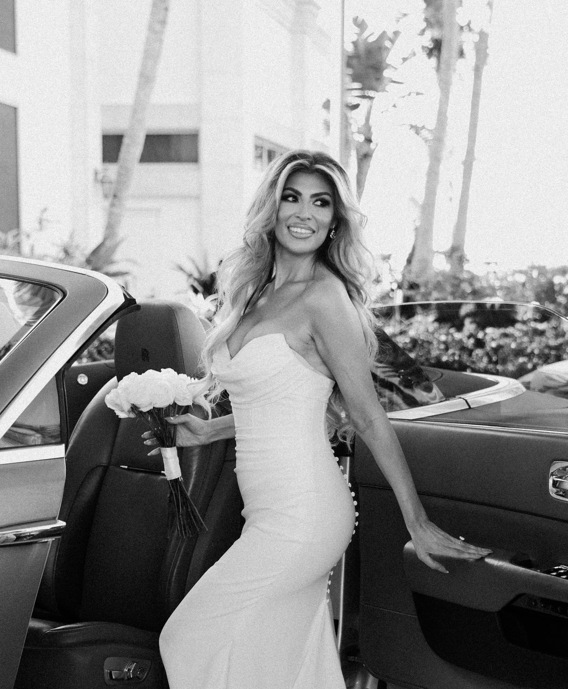 A woman in a white dress holding a bouquet of flowers standing next to a luxury car, with palm trees in the background.