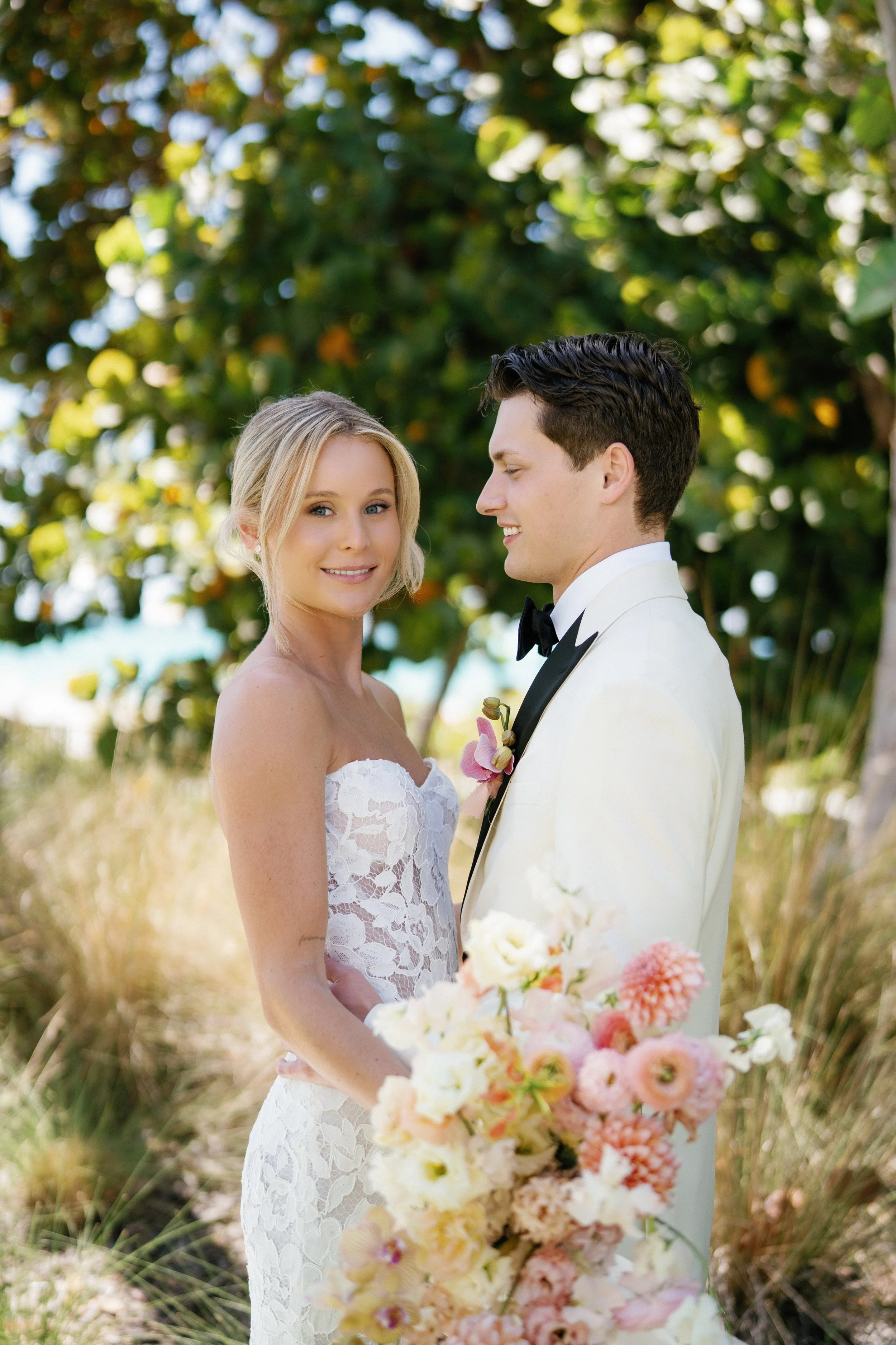 A bride and groom standing outdoors on their wedding day, surrounded by greenery and holding a bouquet of pastel-colored flowers.