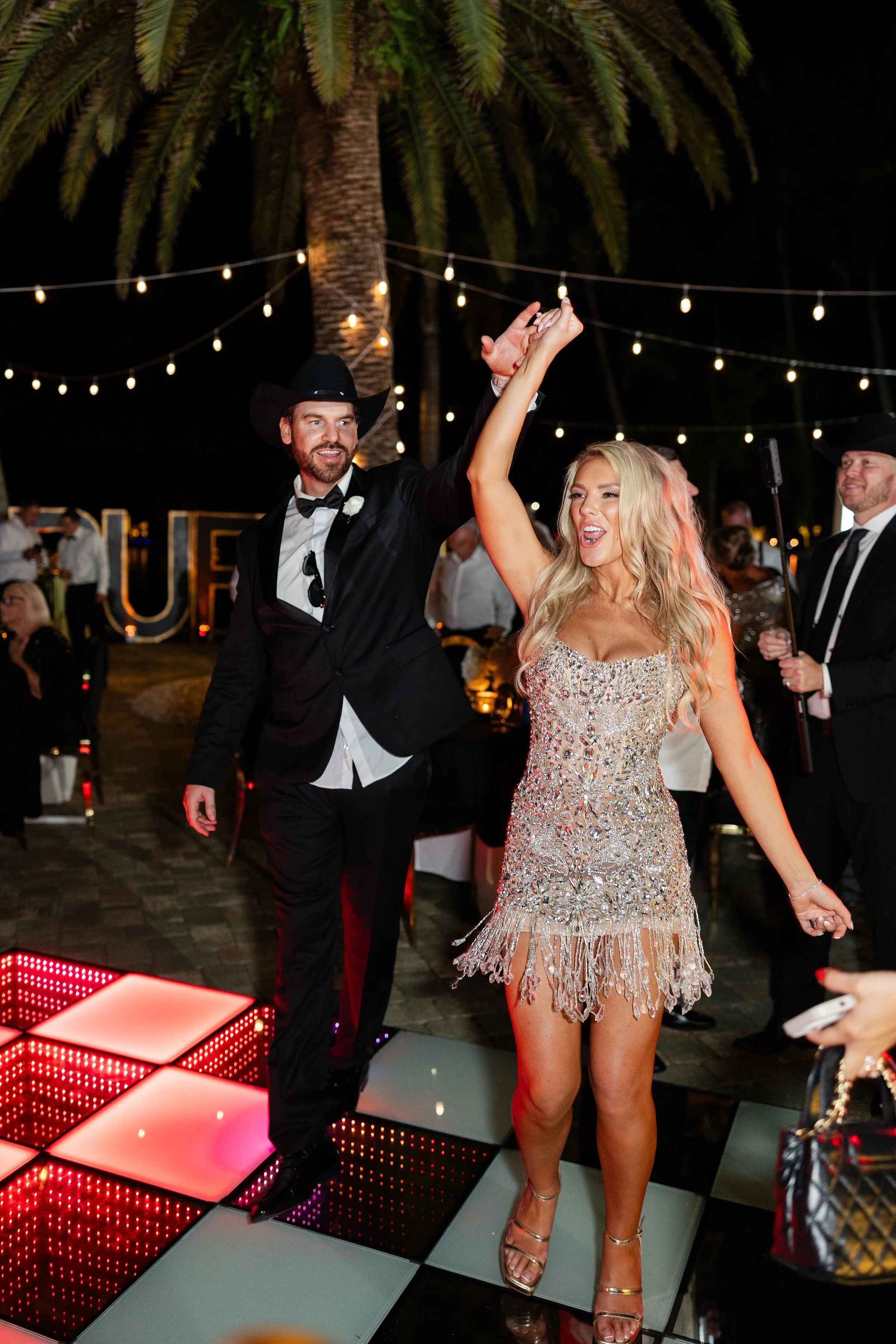 A man dressed in a tuxedo with a cowboy hat and a woman in a sequined dress are dancing at an outdoor event at night with string lights overhead. Others are visible in the background.