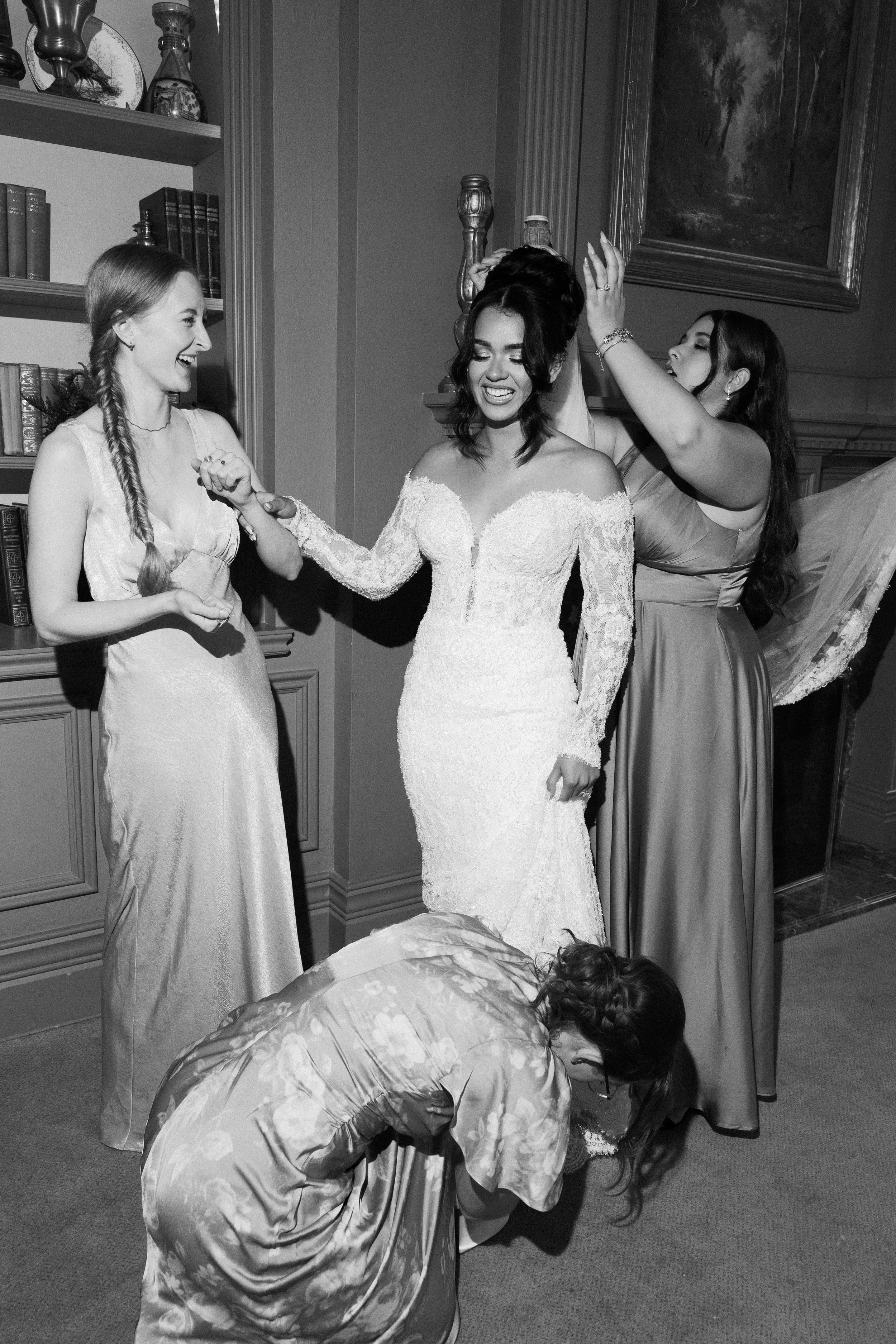 Three women, one in a wedding dress, are celebrating with a cake. One woman is standing and smiling, while another is crouched down, possibly cutting the cake or partaking in a wedding ritual, with a woman in a long dress placing a hand on the bride'