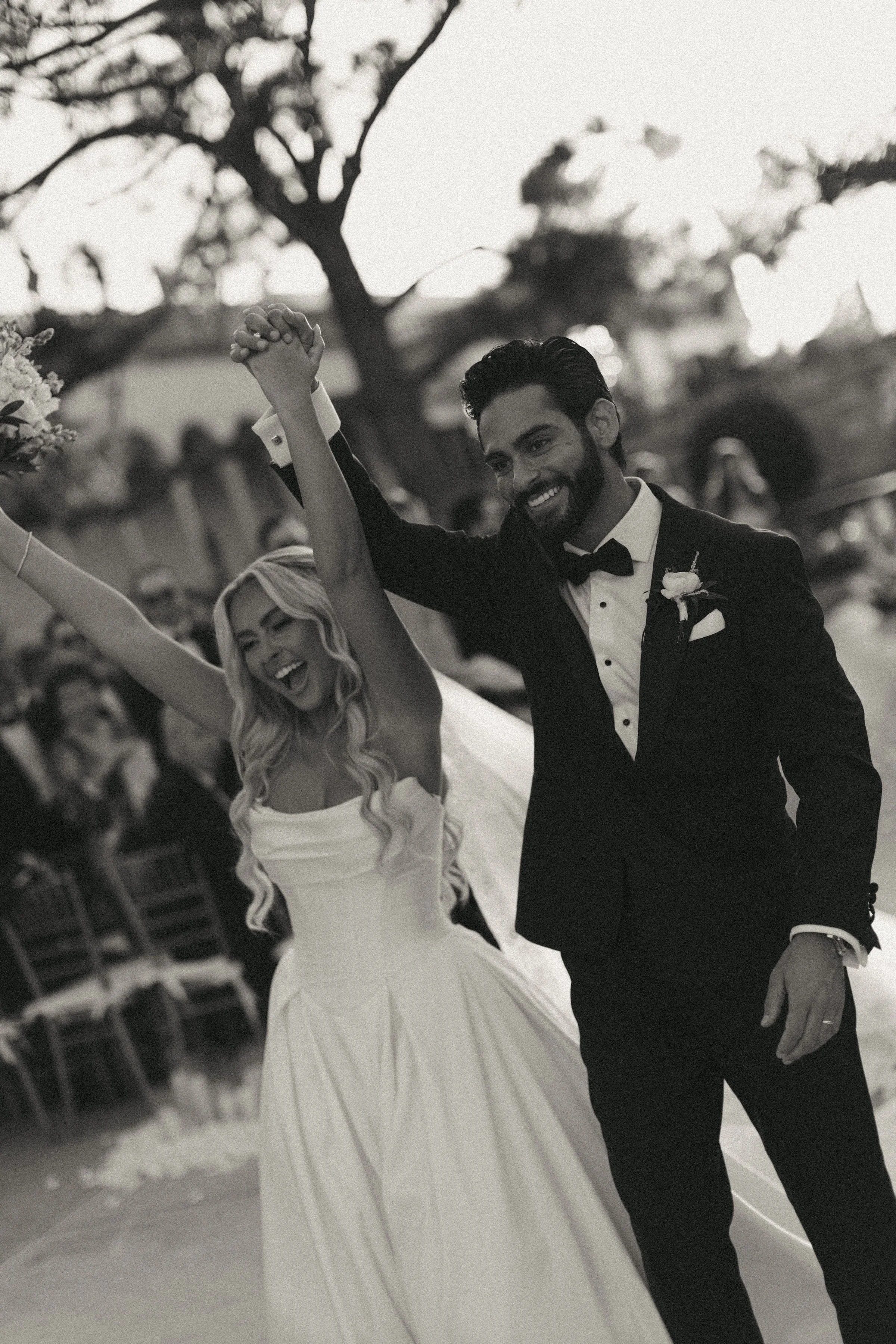 A smiling bride and groom dancing at their wedding, holding hands and raising their arms, outdoors with trees and chairs in the background.