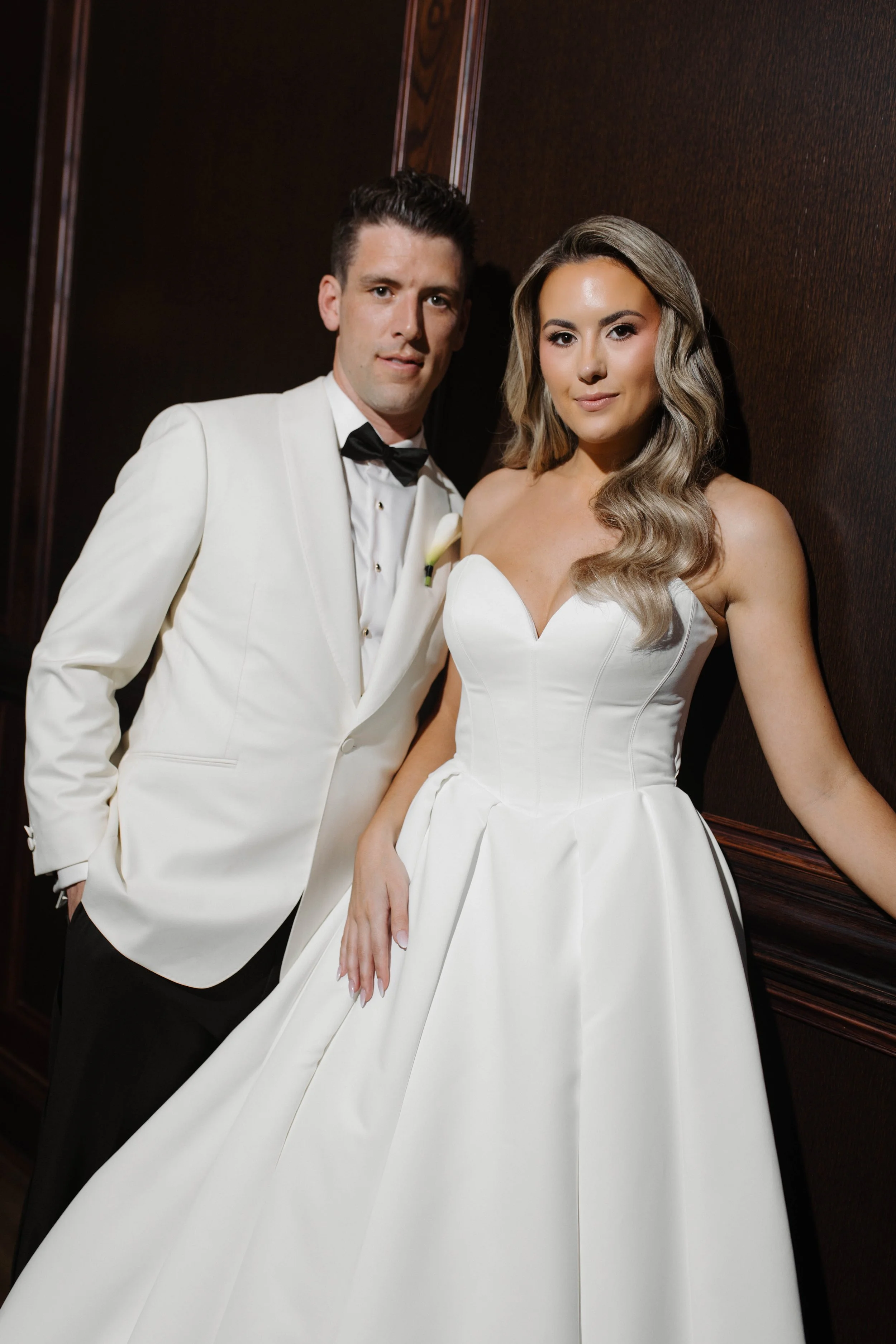 A man and woman dressed in formal wedding attire, standing close together against a dark wood wall.