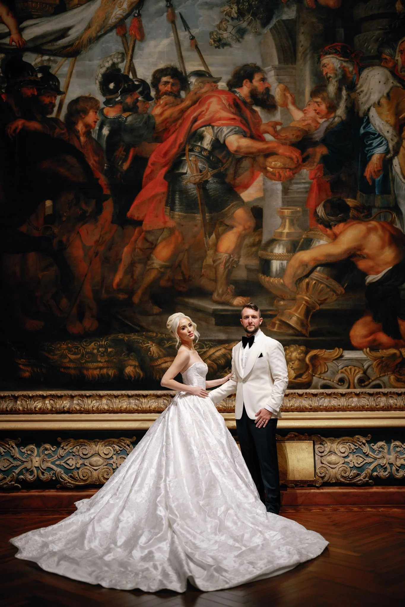 A bride and groom in wedding attire standing in front of a large, ornate painting depicting a historical or mythological scene. The bride is wearing a white wedding gown with a long train, and the groom is in a black tuxedo with a white jacket. They 