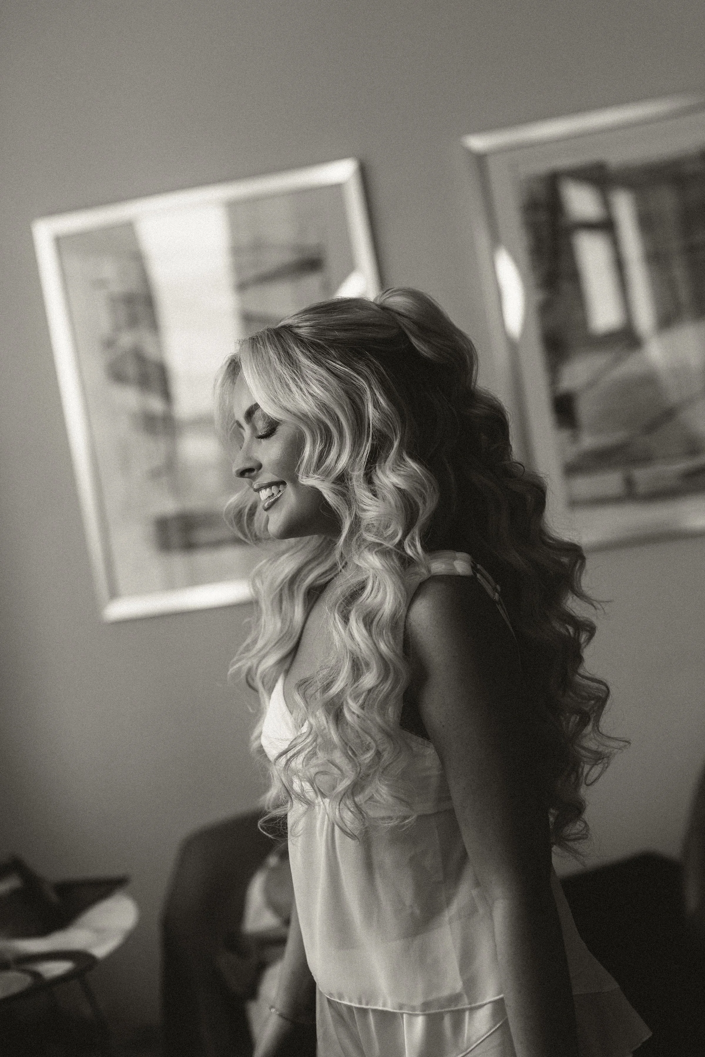 A woman with long, wavy, blonde hair styled in loose curls, smiling with her eyes closed, standing indoors with framed pictures on the wall behind her.