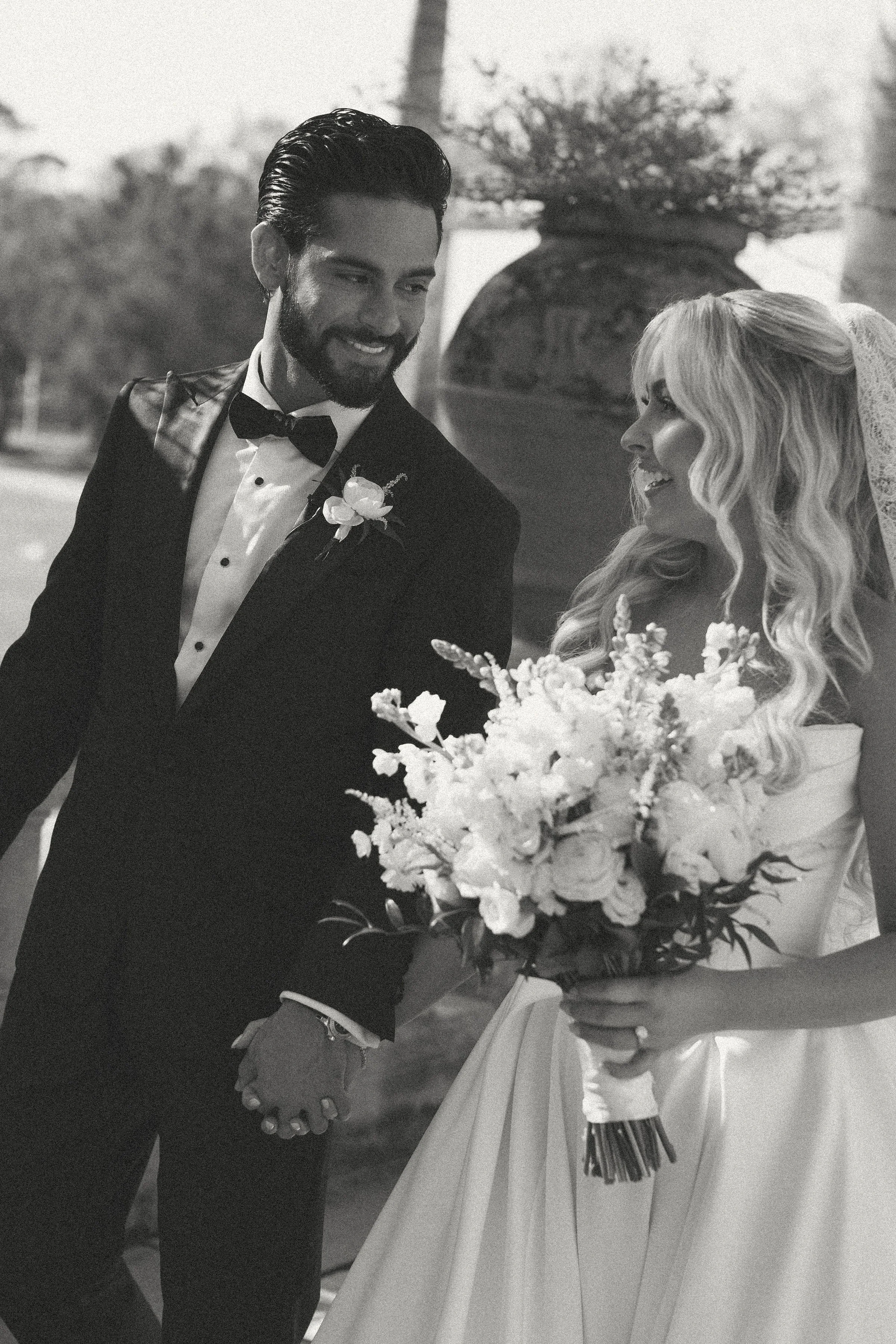 A black and white photo of a groom and bride holding hands, with the groom smiling and wearing a tuxedo, and the bride holding a large bouquet of flowers.