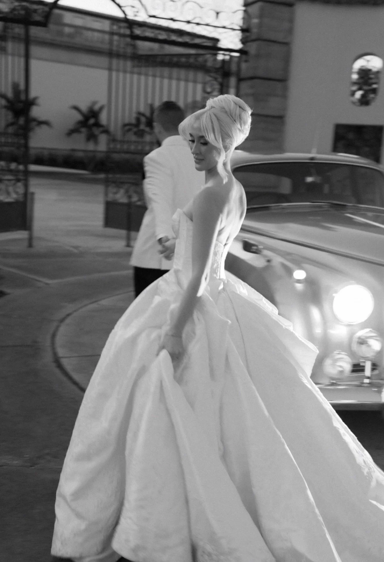A woman in a wedding dress walking beside a man in a suit, with a vintage car in the background, in a black and white photo.