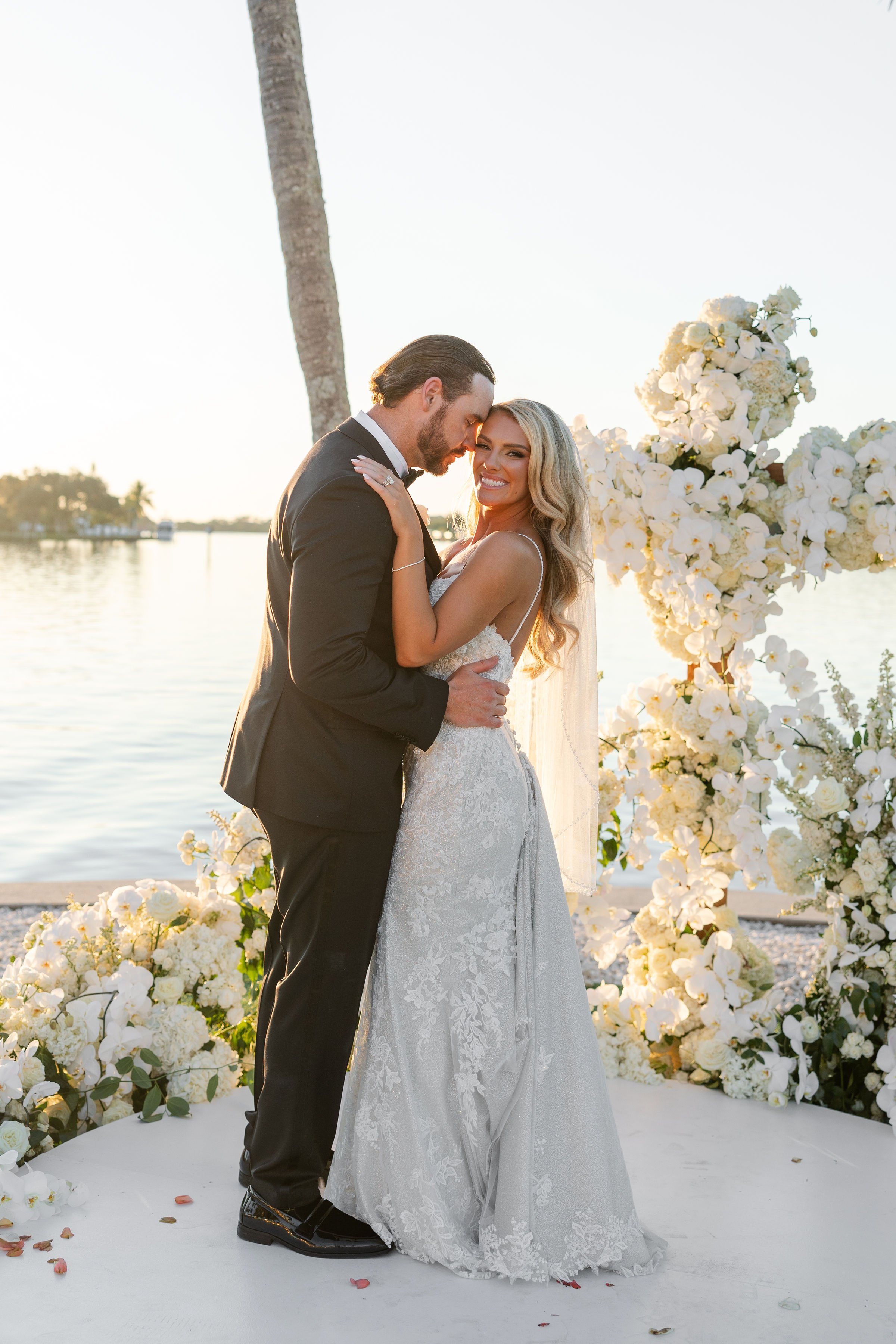 A bride and groom share a moment during their wedding ceremony by the water, with a backdrop of white flowers and a tall palm tree, during sunset.