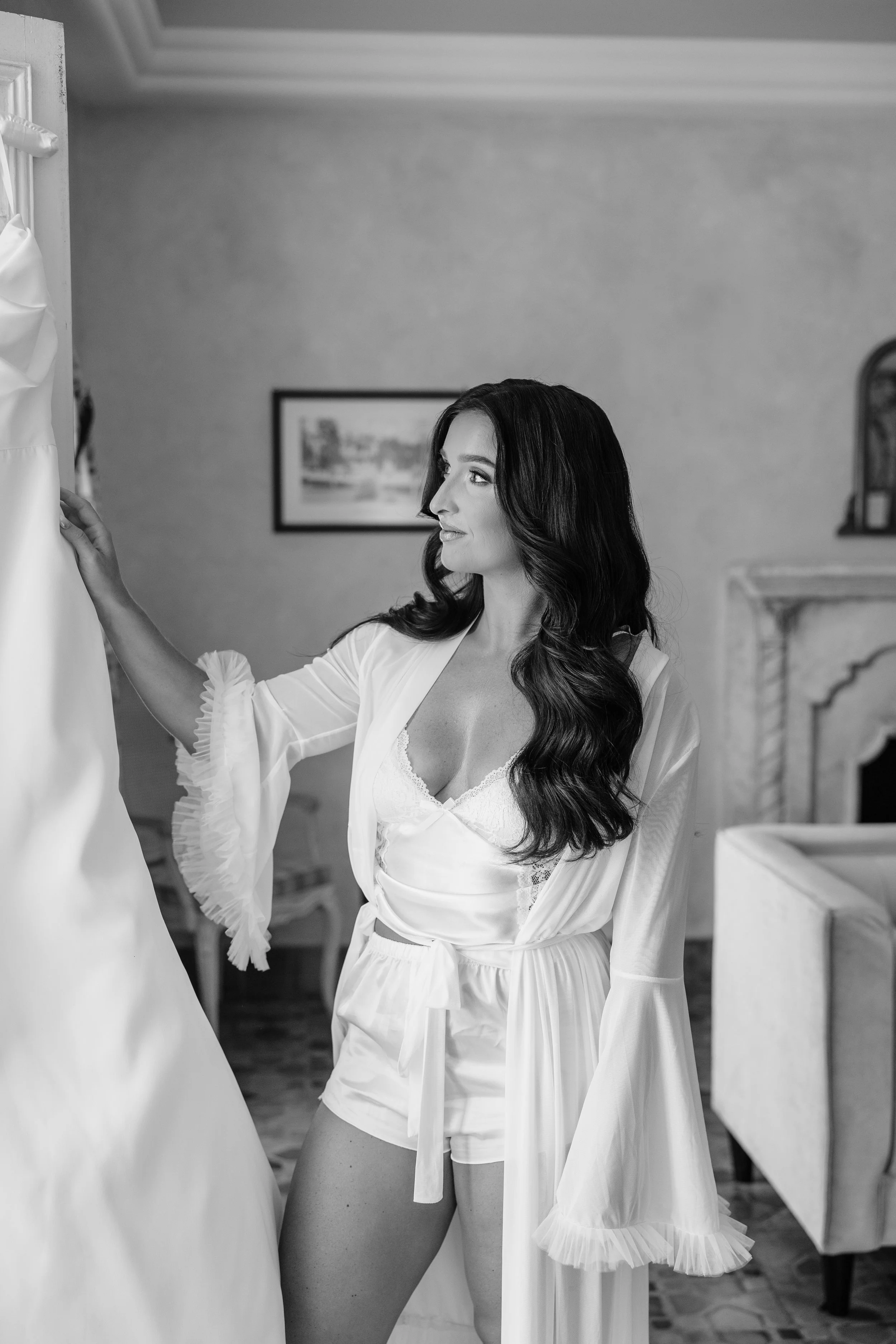 A woman with long dark wavy hair stands indoors, looking at a white satin dress hanging on a wall or mannequin.