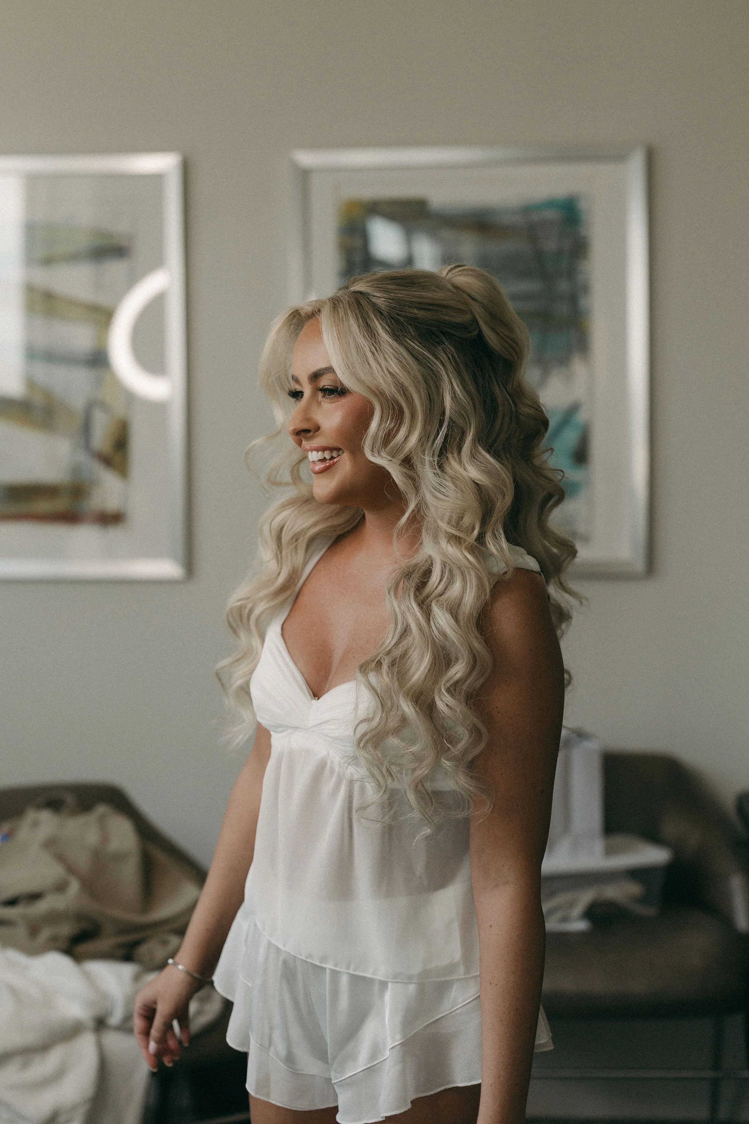 A woman with long, wavy blonde hair smiling in a white dress, standing in a bedroom with unmade bed and three framed abstract art pieces on the wall.