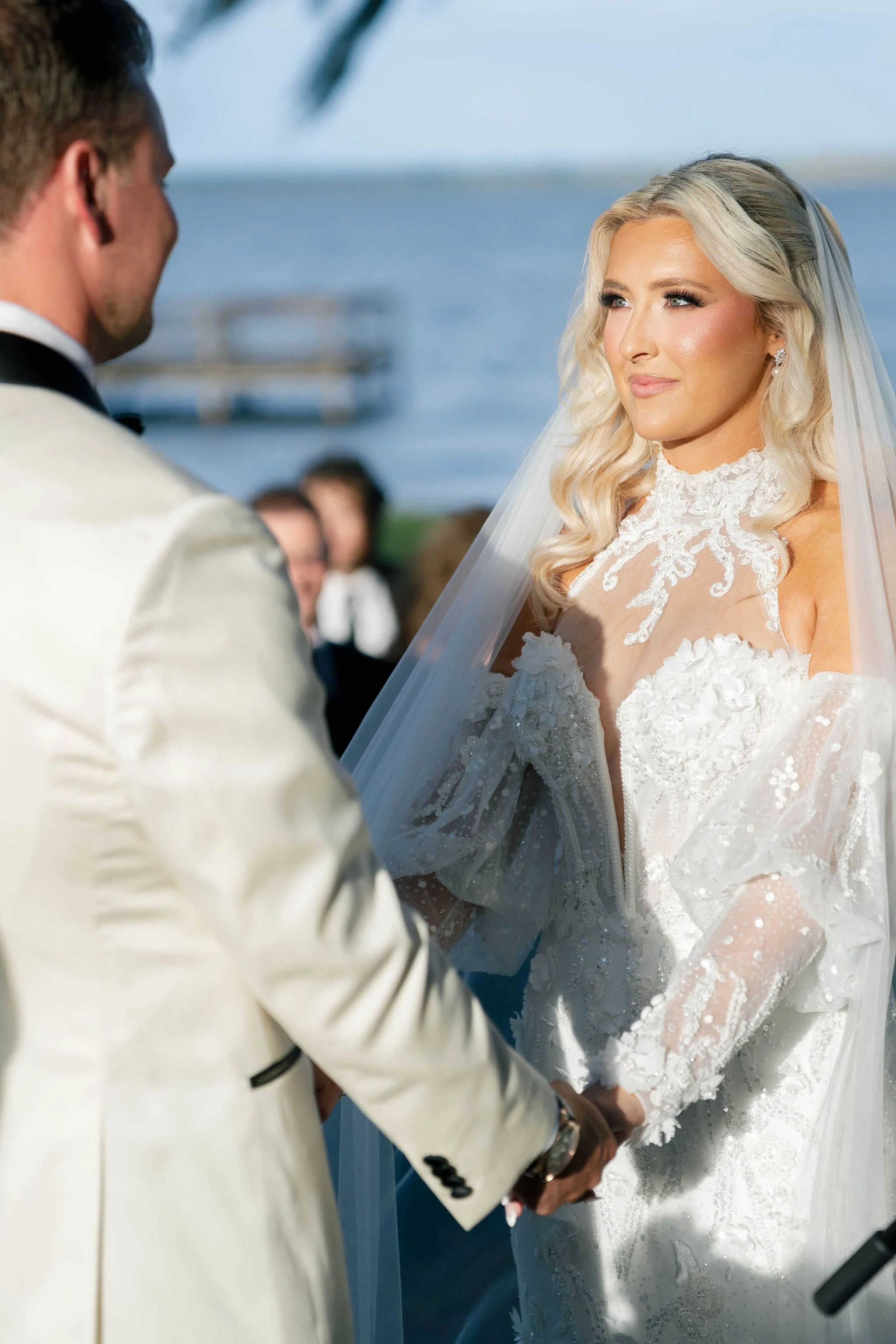 A bride and groom hold hands during their wedding ceremony outdoors near the water, with the bride wearing a white lace gown and the groom in a cream-colored suit.