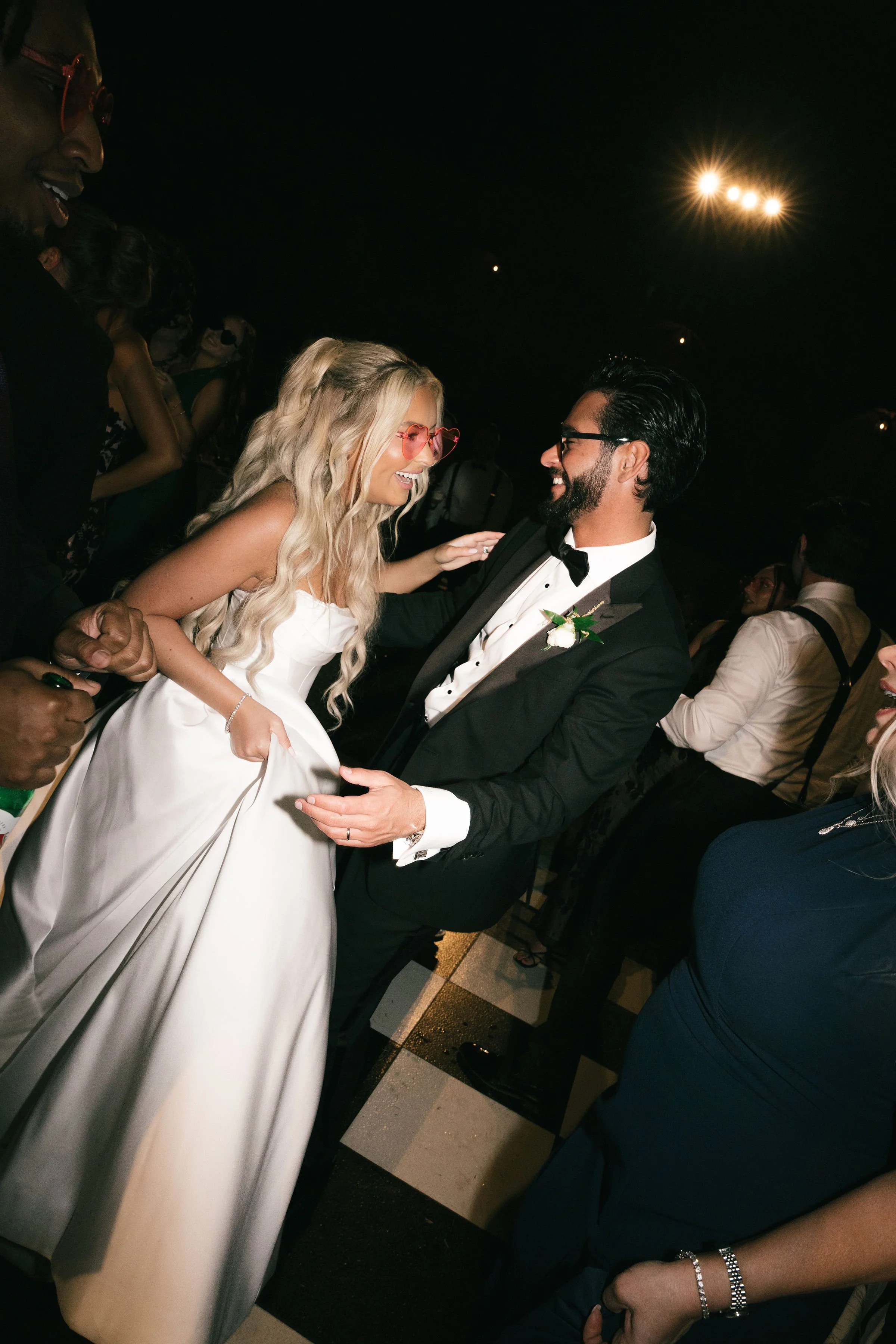 A bride and groom are dancing at their wedding reception. The bride is wearing a white gown and pink heart-shaped sunglasses, and the groom is wearing a black tuxedo with a white shirt and black bow tie. They are smiling and holding hands, surrounded
