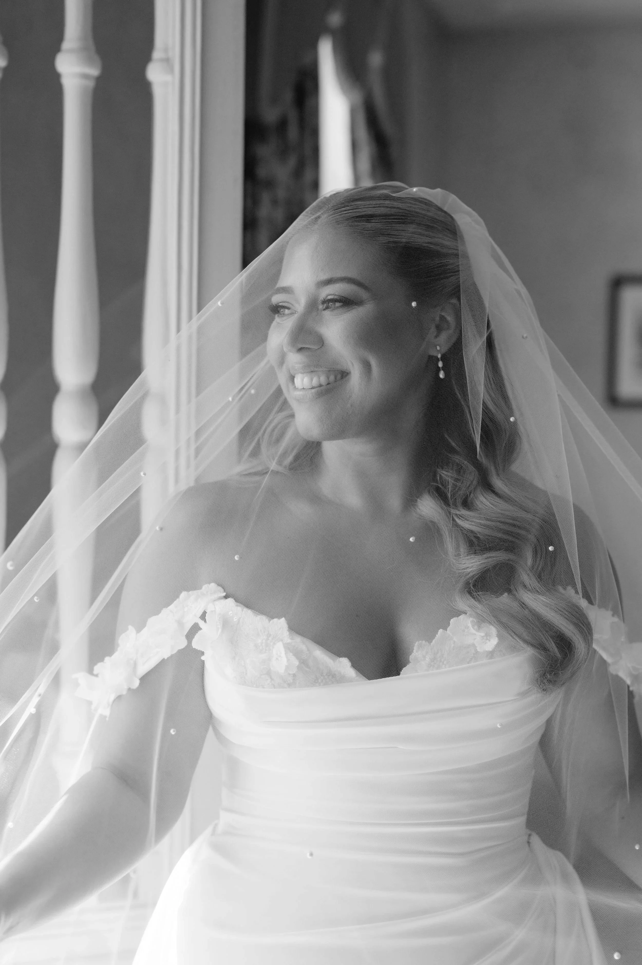 Black and white photo of a smiling bride in a wedding dress and veil, standing in front of a mirror.