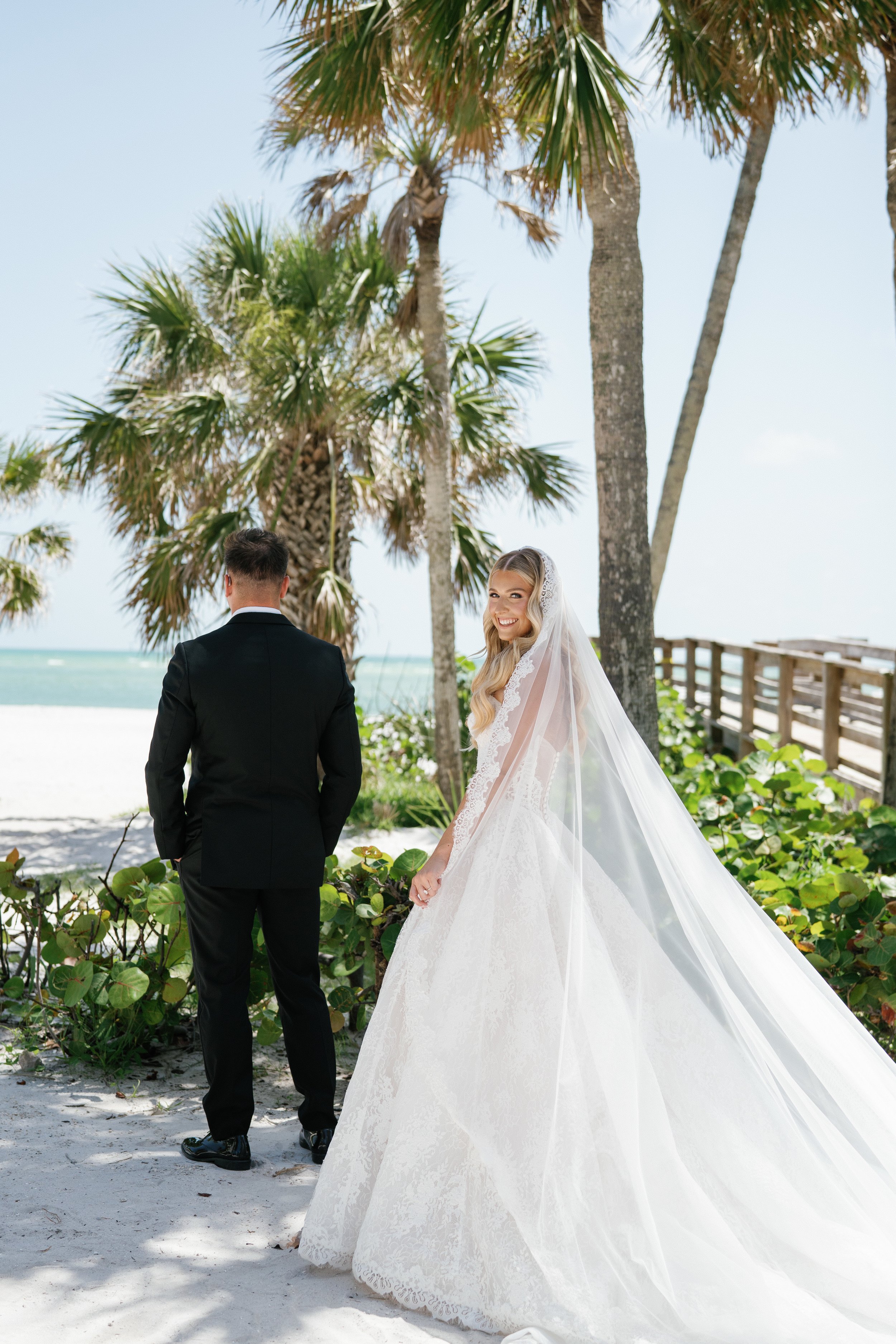 A bride in a white wedding gown smiling while standing on a beach with palm trees, a wooden railing, and sand, facing a groom in a black tuxedo.
