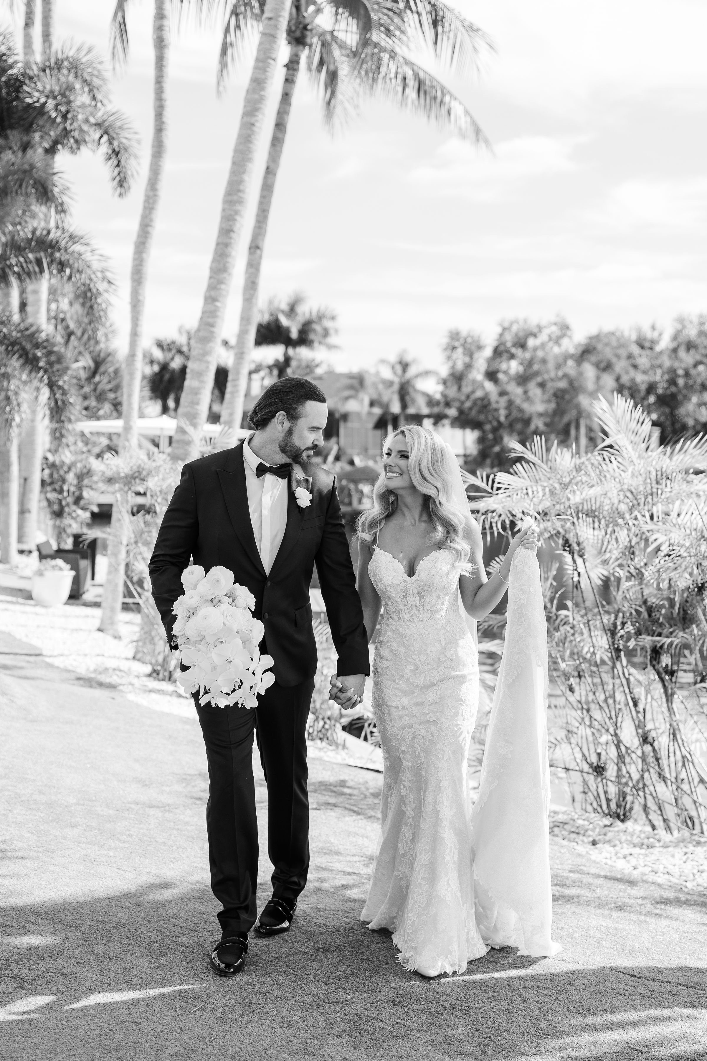A bride and groom walking hand in hand outdoors on a sunny day. The groom is dressed in a tuxedo, holding a bouquet of white flowers, and the bride is wearing a lace wedding dress with a veil. Palm trees and lush greenery are in the background.