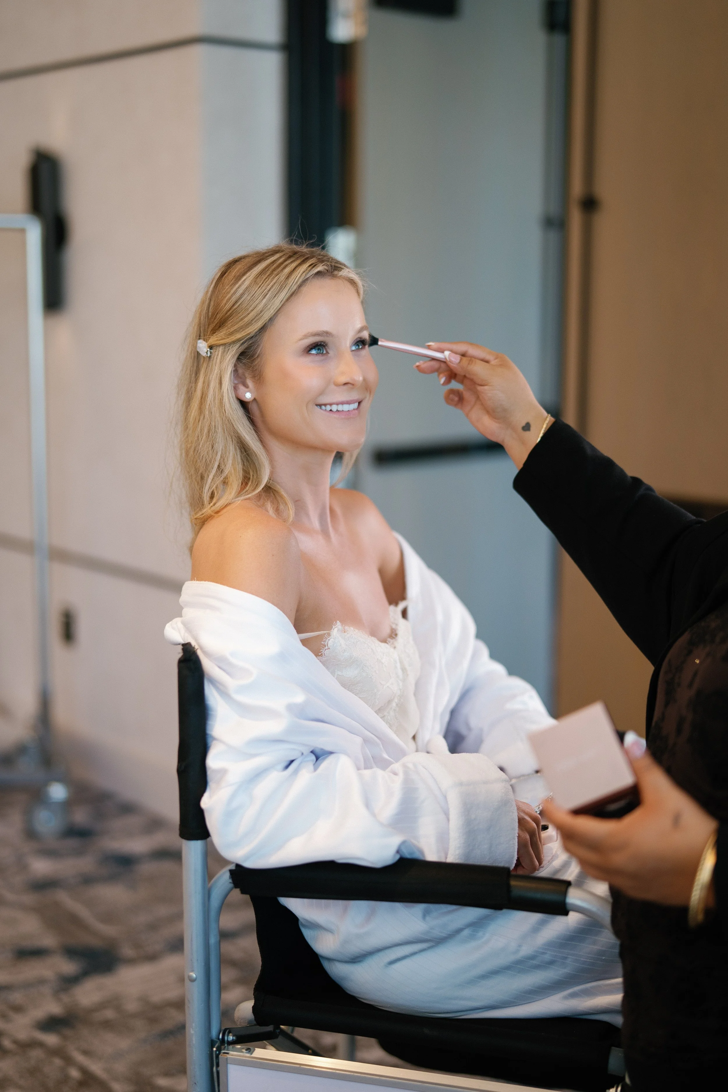 A woman with blonde hair and a white off-shoulder top smiling as someone applies makeup to her face with a brush. She is sitting in a wheelchair, and there is another person holding a palette in front of her.