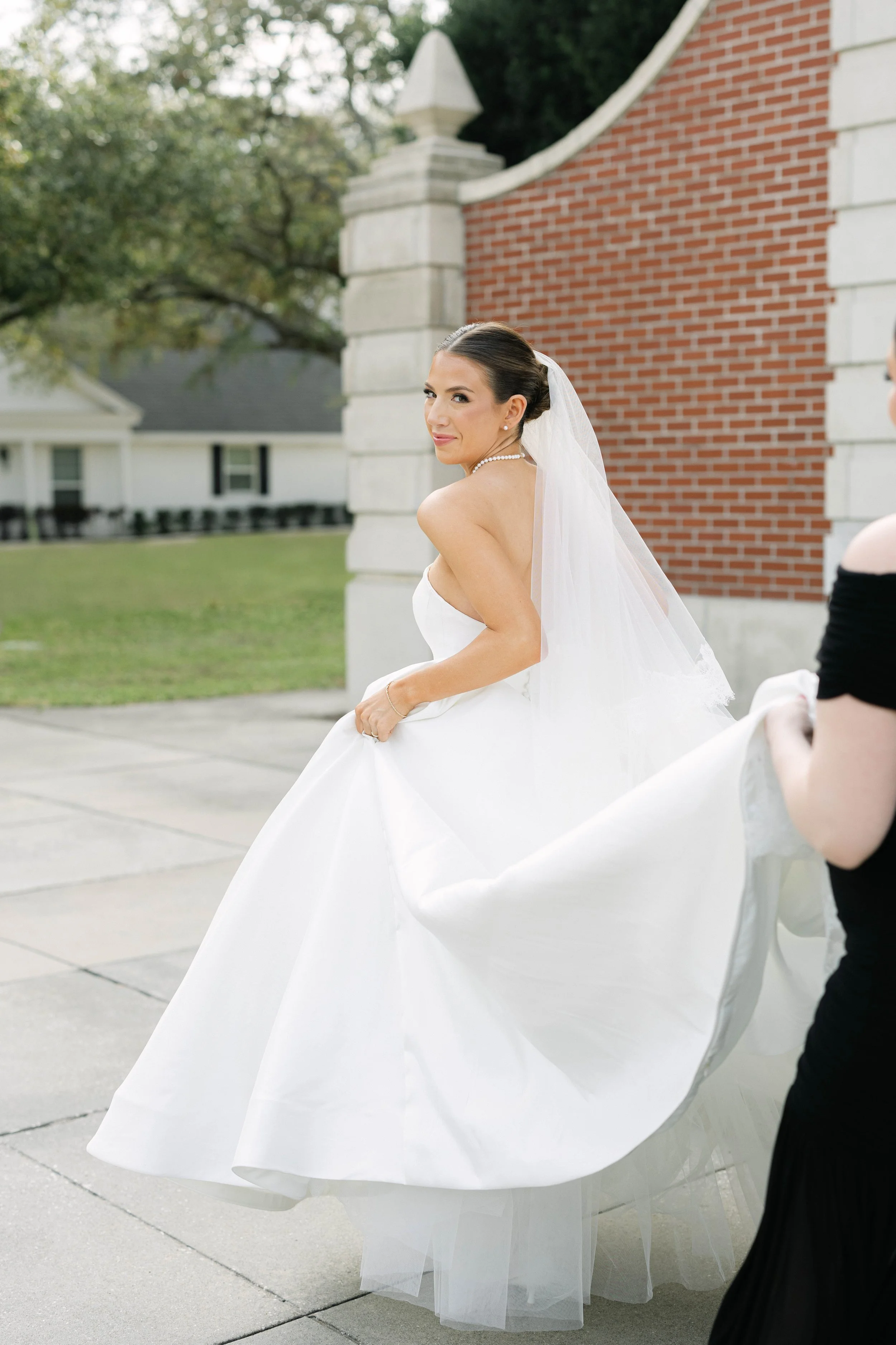 A bride in a wedding dress with a veil and jewelry, holding the hem of her dress, standing outdoors in front of a brick and stone building with trees in the background, smiling and looking over her shoulder.