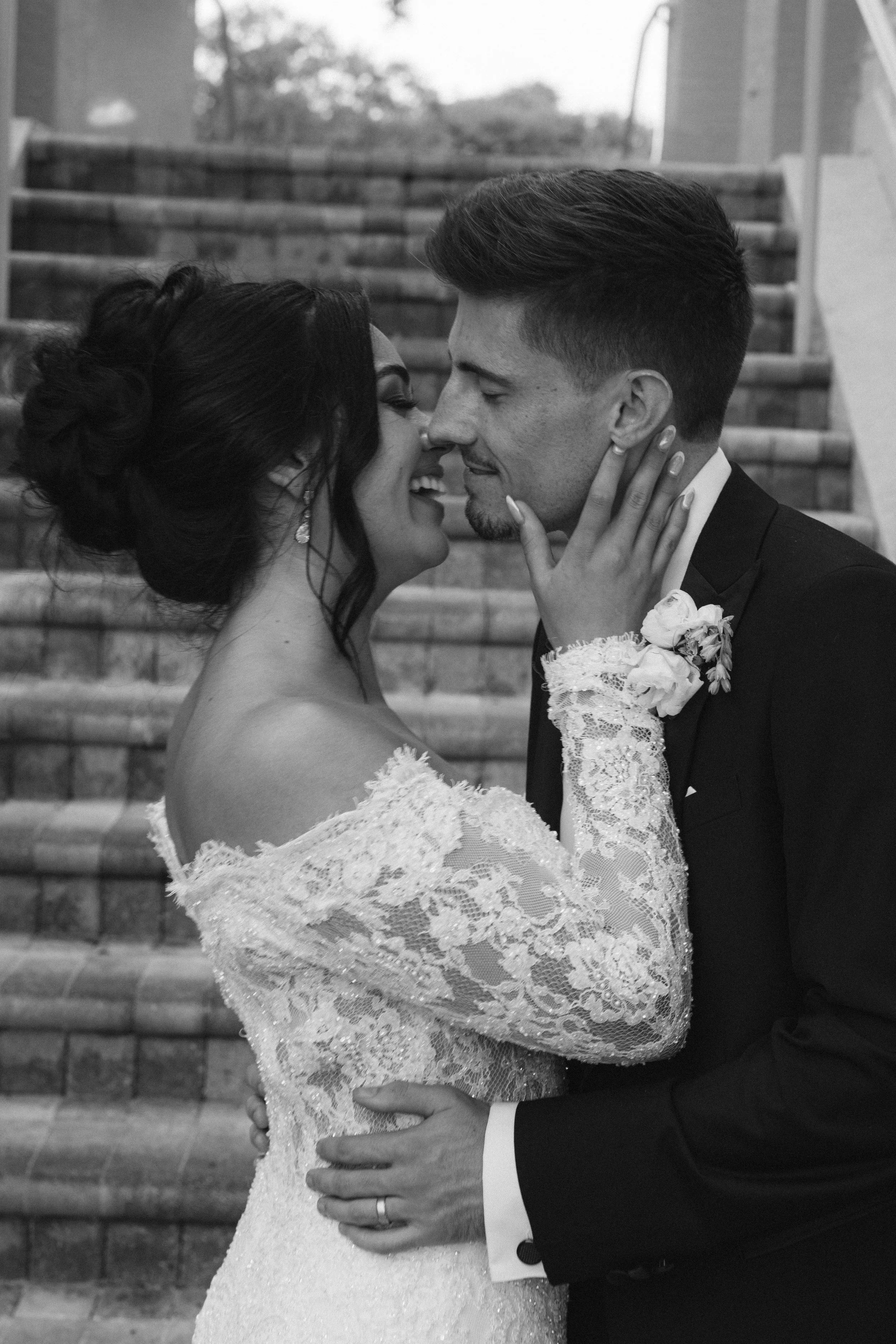 A bride and groom are close together, about to kiss, on steps outside. The bride is wearing a lace wedding gown and lace glove, and the groom is in a tuxedo with a boutonniere.