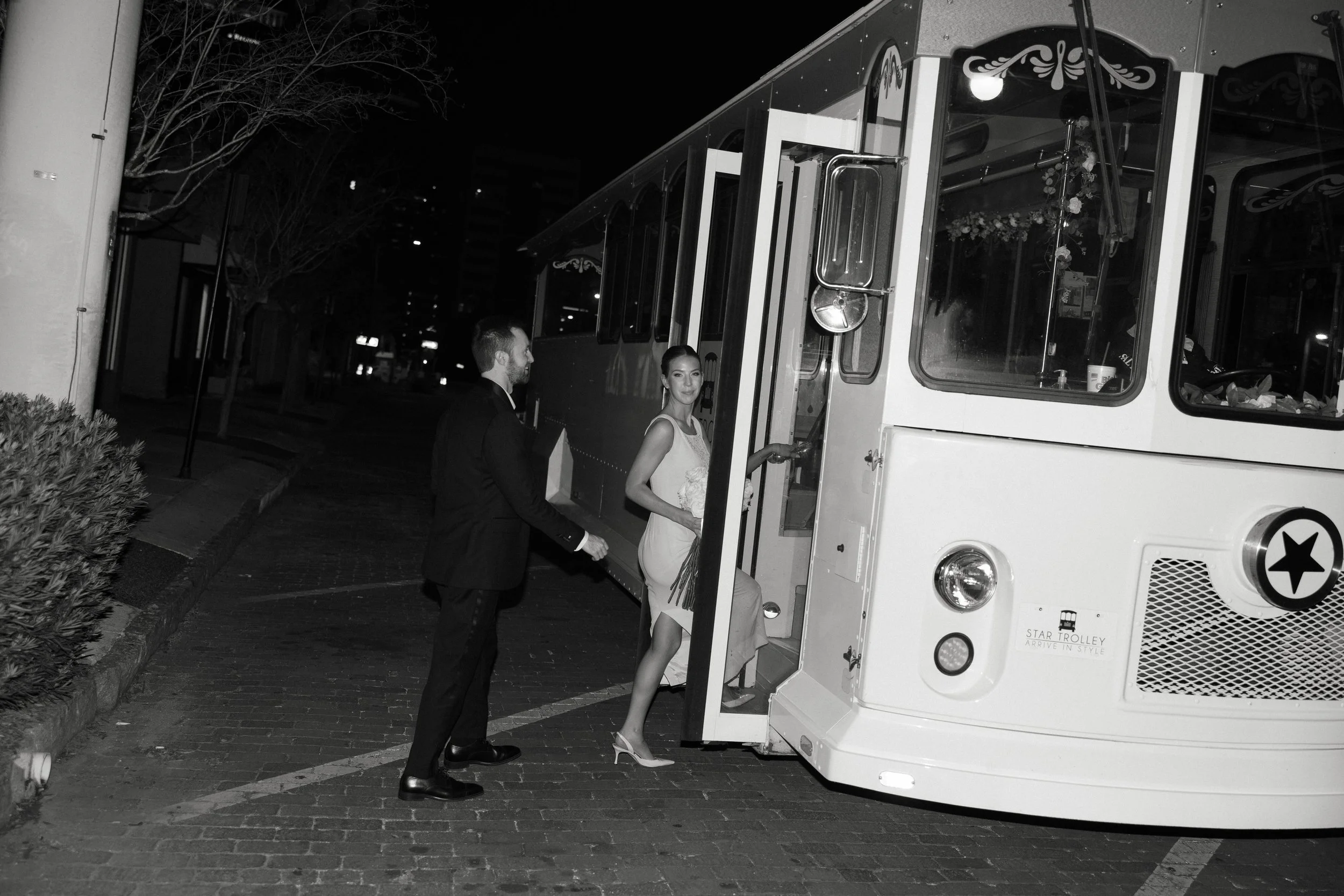 A bride and groom boarding a vintage-style trolley at night. The bride is holding a bouquet of flowers and the groom is dressed in a suit. The trolley has decorative details and the scene is outdoors with trees and buildings in the background.