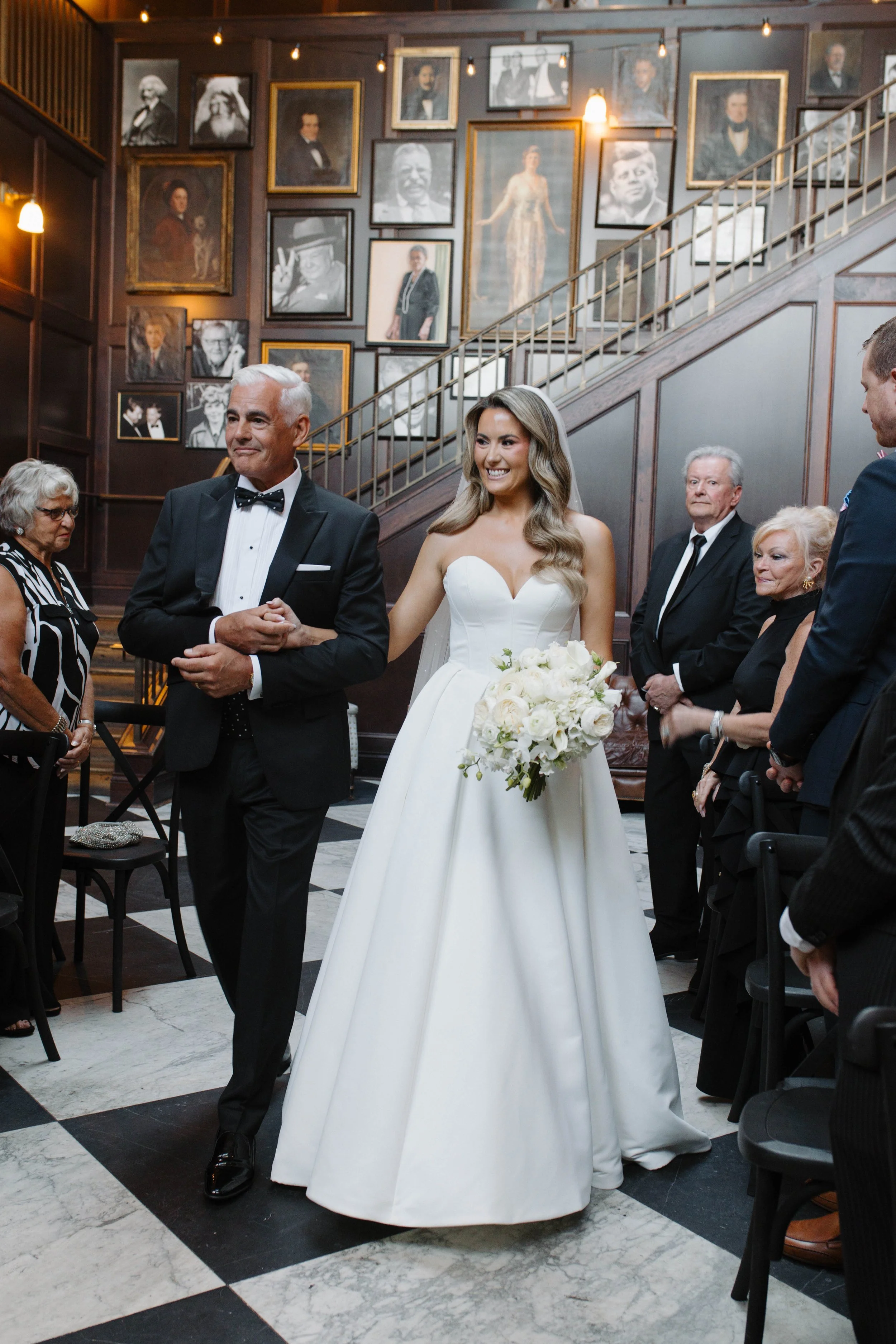 A bride in a white wedding gown celebrates with her father, who is dressed in a black tuxedo, as family members stand around in an elegant wood-paneled room with framed black-and-white photographs on the wall at Oxford exchange in Tampa fl . 