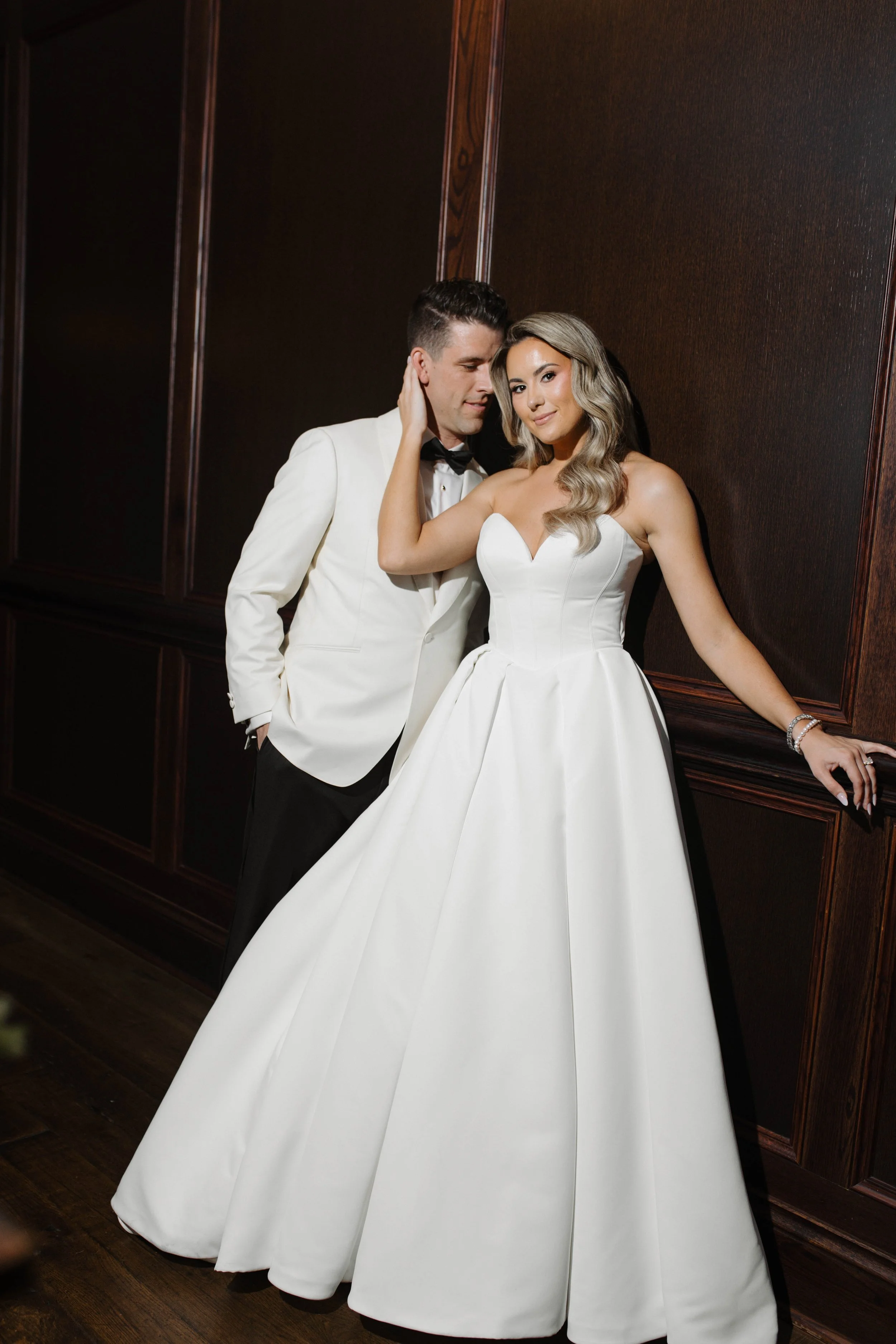 A bride in a strapless white wedding gown with a full skirt and a groom in a white tuxedo with black bow tie, standing close together against a dark wood-paneled wall.