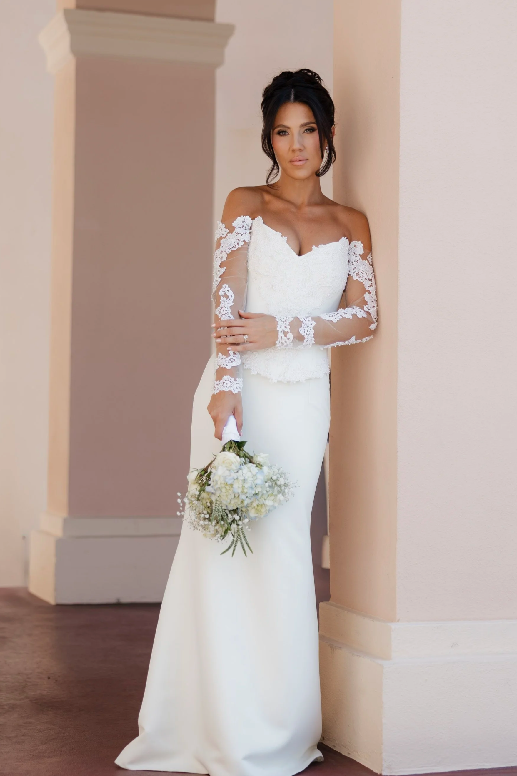 A woman in a white wedding dress with lace sleeves, holding a bouquet of white flowers, standing against a beige wall with architectural details.