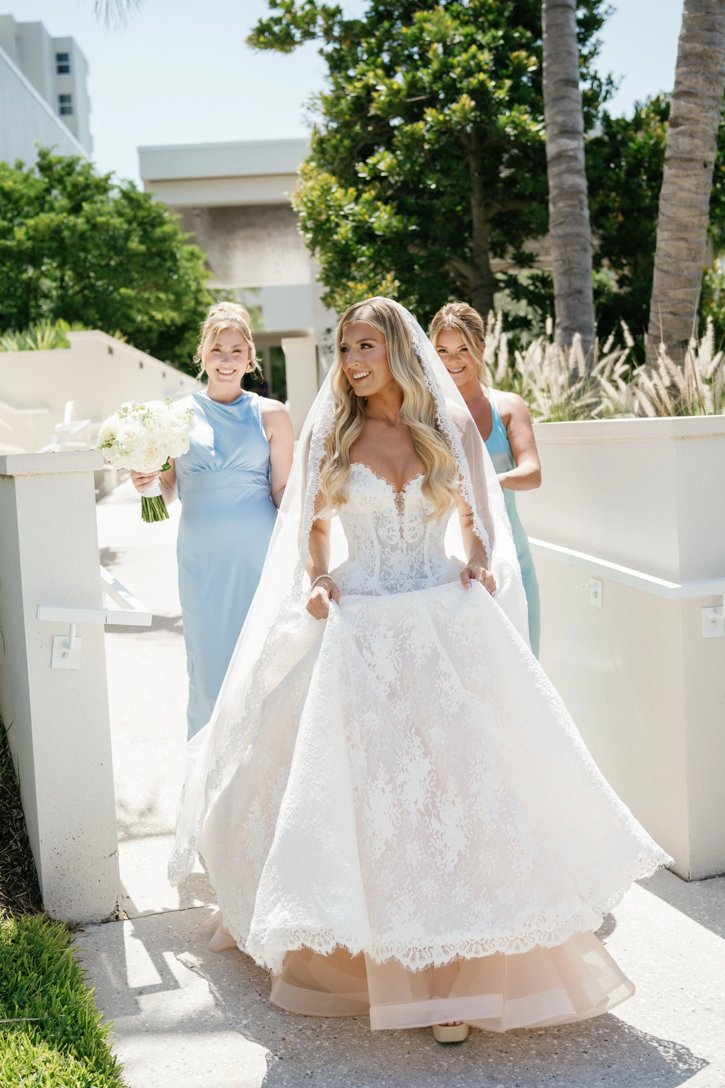 Bride in a white lace wedding gown with a v-neckline and veil, holding the skirt of her dress, walking outdoors on a sunny day, with two bridesmaids in light blue dresses, and smiling. There are palm trees and greenery in the background.