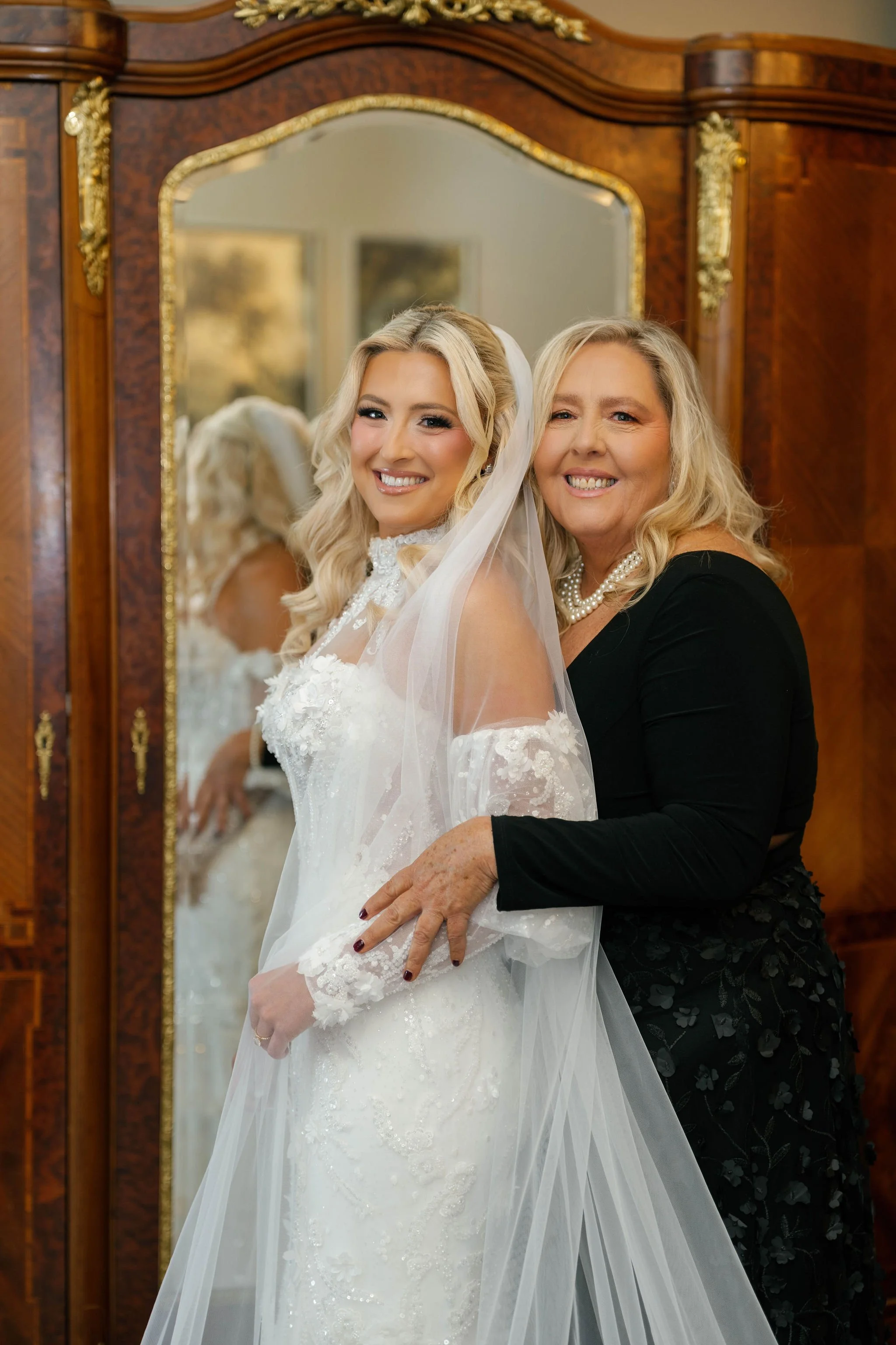A bride in a white wedding dress with lace details, smiling, standing in front of a large mirror, with a woman in black and pearl necklace beside her, both smiling. The background features a wooden antique wardrobe with gold accents.